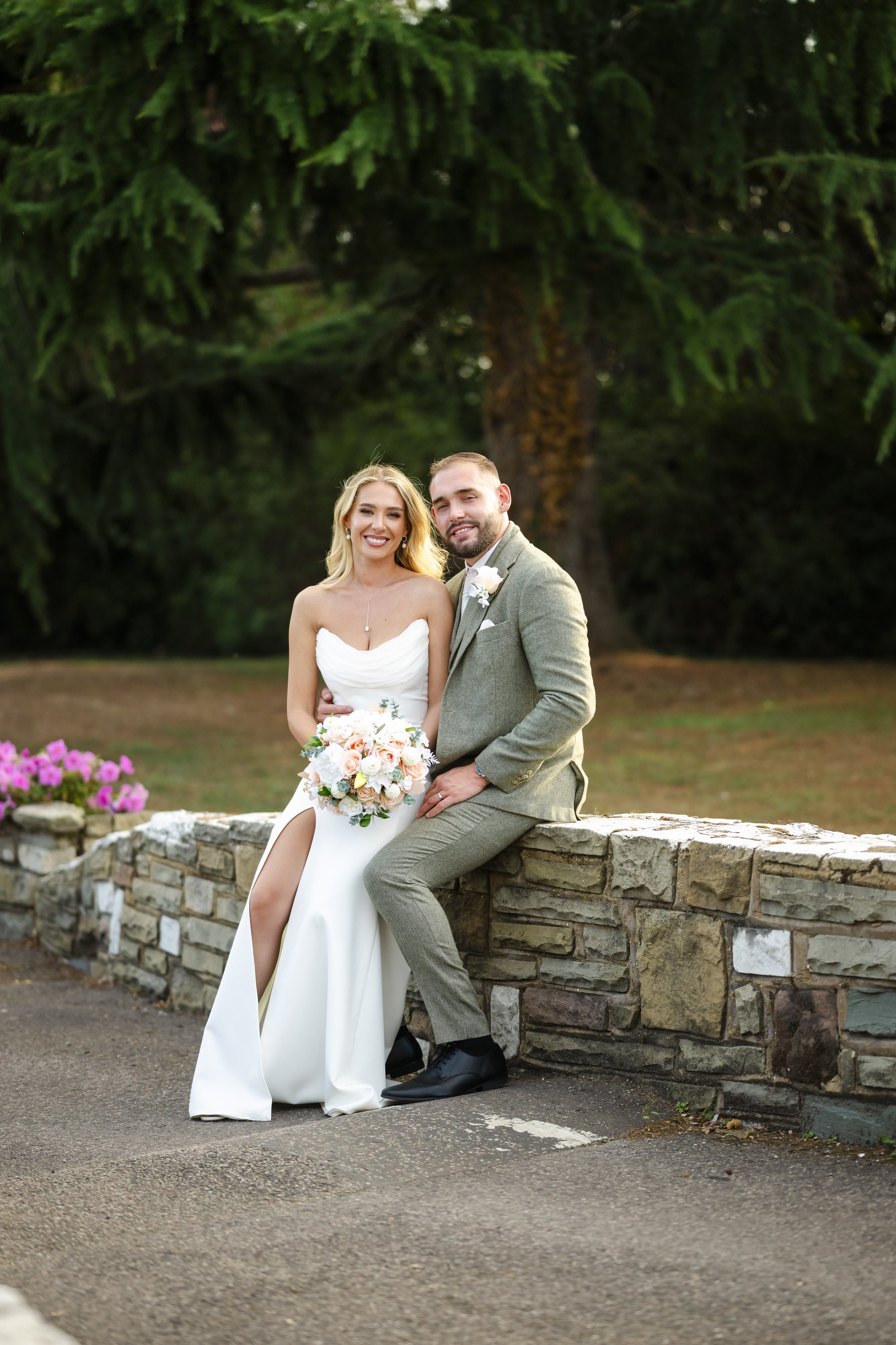 A bride and groom sitting on a stone wall outdoors, smiling, with green trees and pink flowers in the background. The bride is wearing a white gown and holding a bouquet, and the groom is in a gray suit.