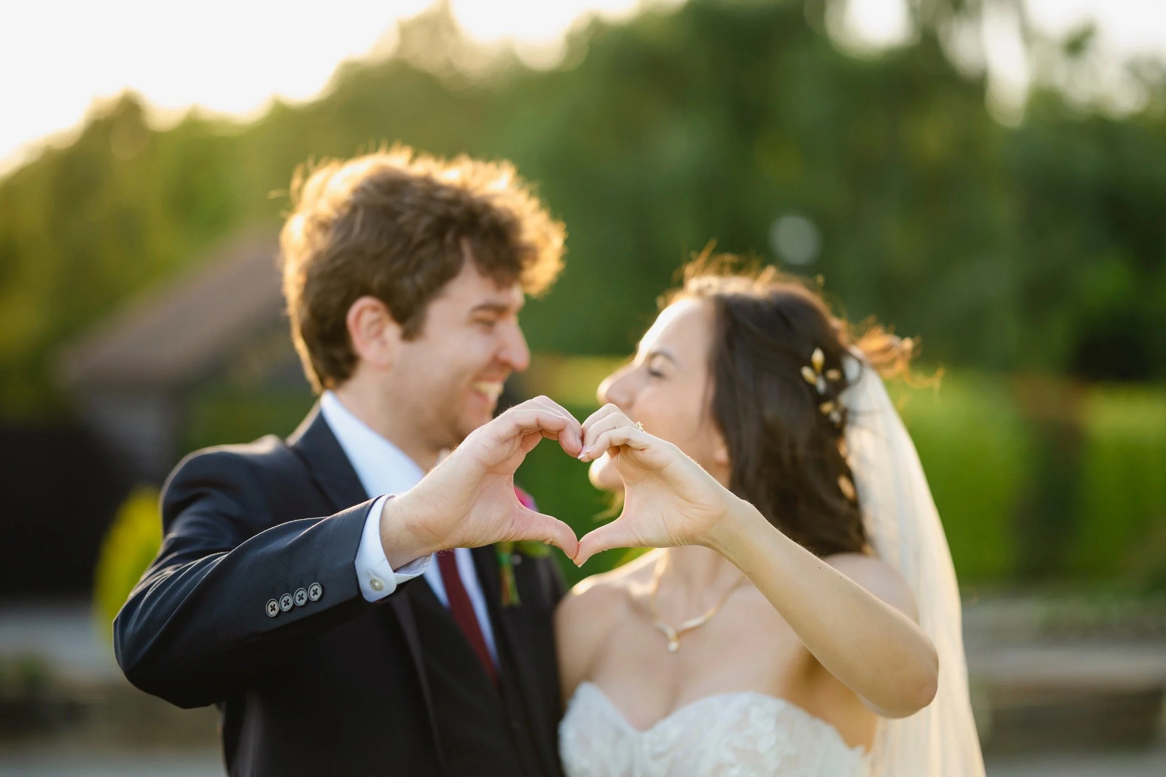A newlywed couple smiling and forming a heart with their hands outdoors during sunset.