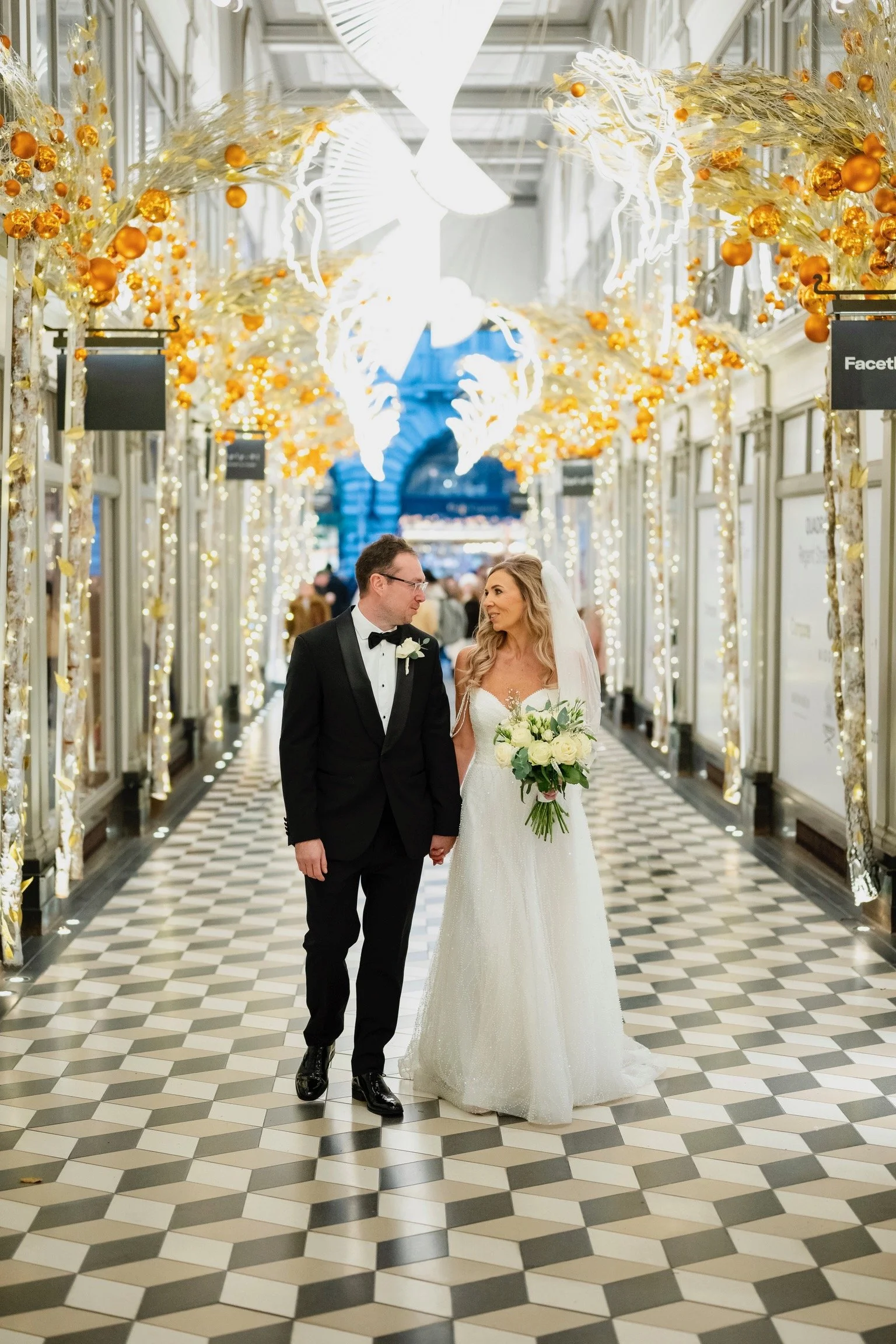 A bride and groom holding hands and walking through a decorated indoor space, with the bride holding a bouquet of white roses and greenery, surrounded by festive lights and holiday decorations.