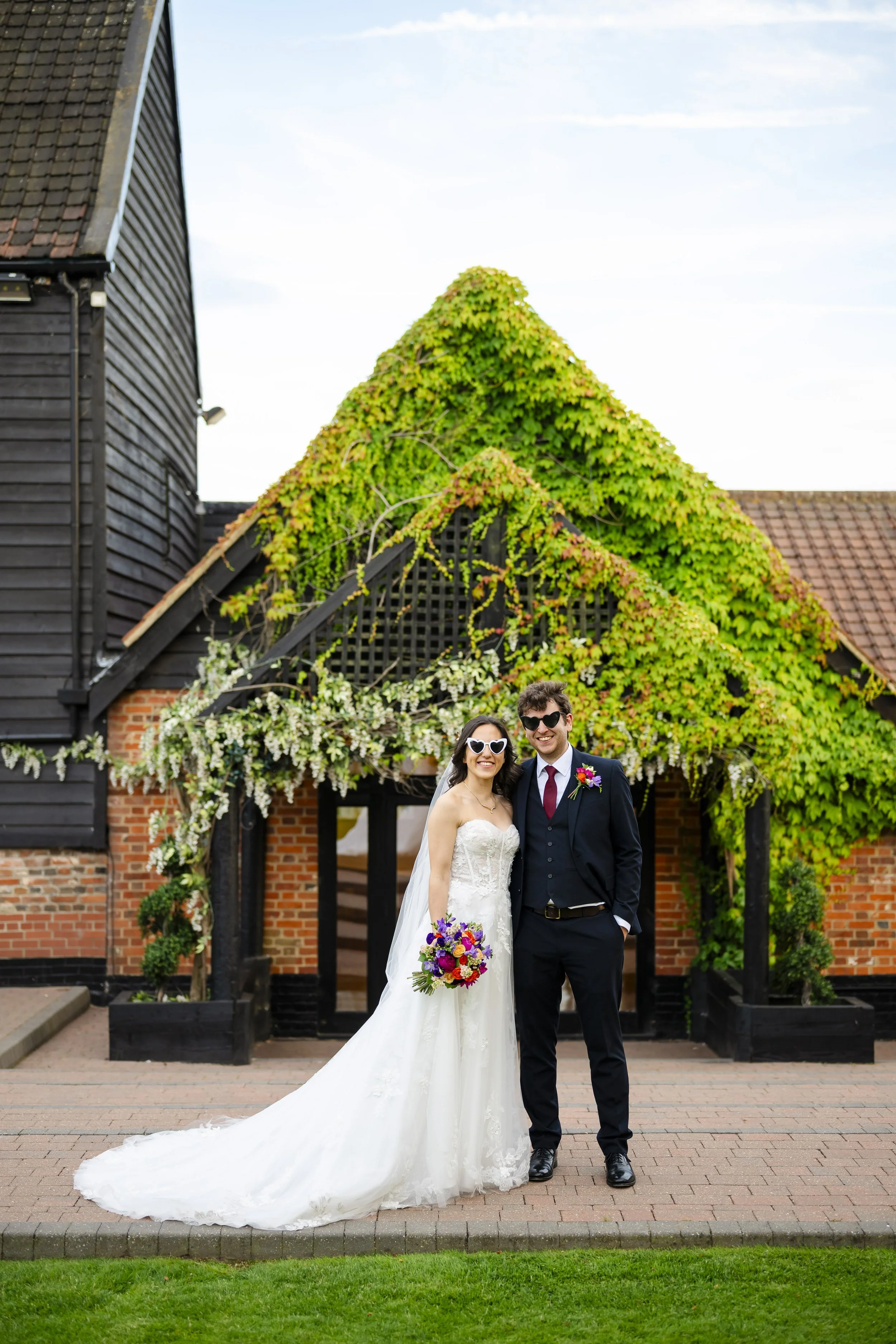 Bride and groom smiling in wedding attire in front of a brick and wooden building with greenery on the roof, holding flowers.