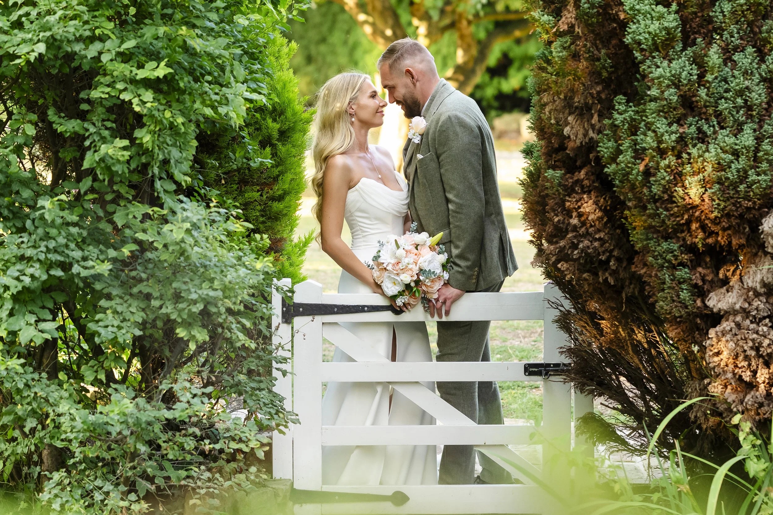 A bride and groom standing close together behind a white picket fence, surrounded by lush greenery, during their wedding.
