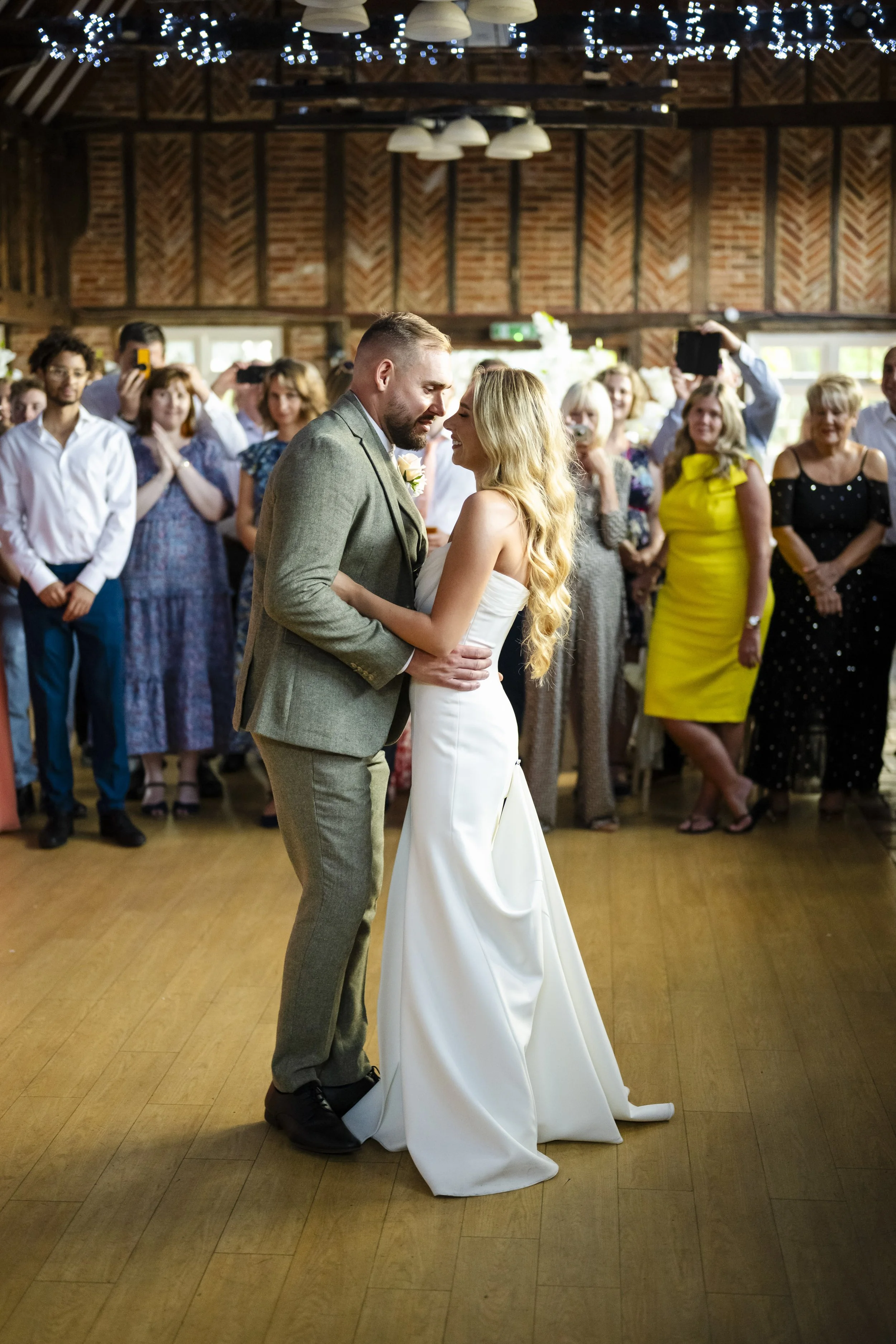 A bride and groom sharing a dance at their wedding reception, surrounded by seated and standing guests in a rustic venue with wooden walls and overhead string lights.
