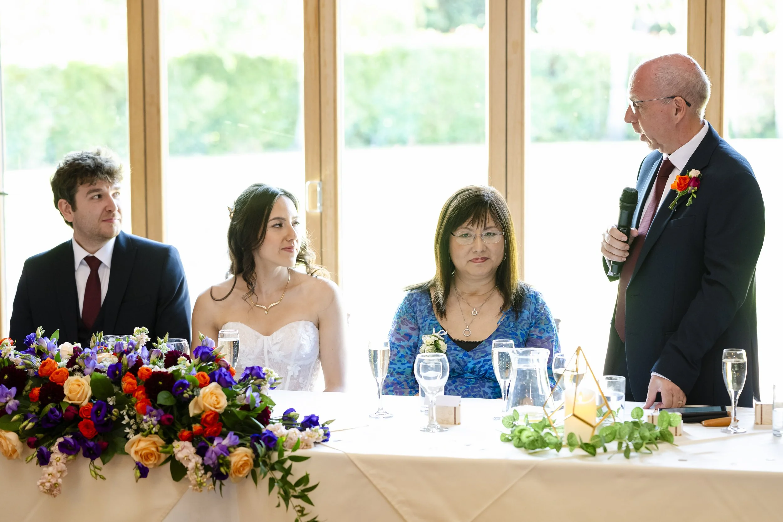 A wedding reception with a man giving a speech while standing next to a table with three seated guests and a flower arrangement.