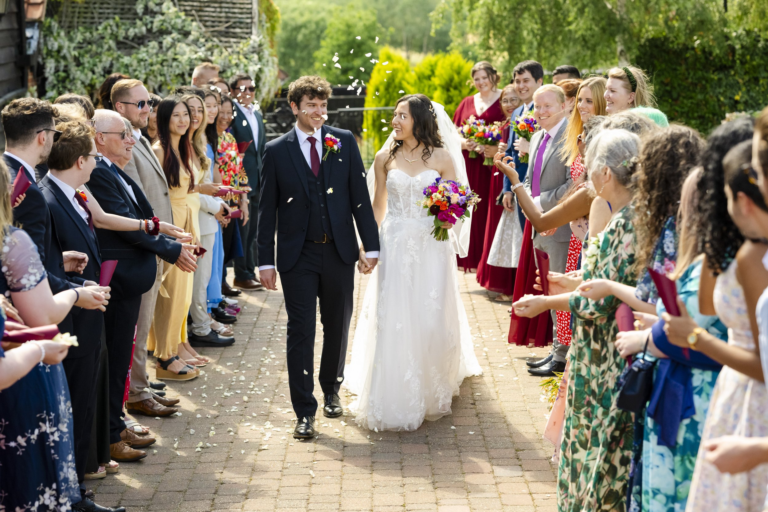 Bride and groom holding hands walking down a garden path, surrounded by smiling wedding guests releasing flower petals, with trees and greenery in the background.