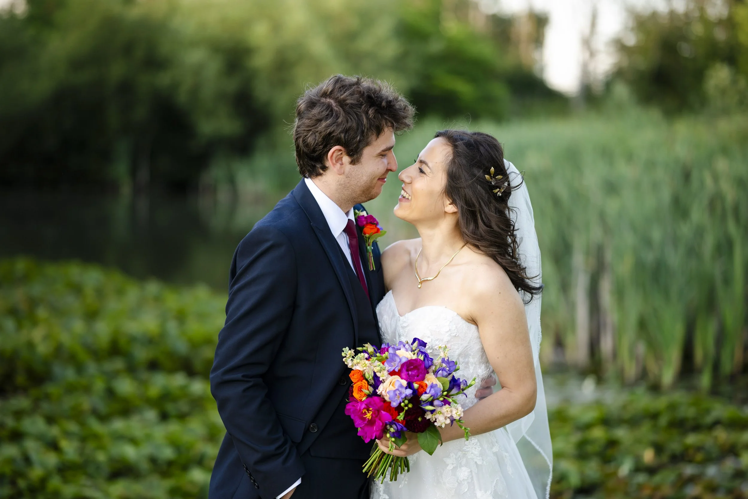 A newlywed couple gazing at each other, standing outdoors by a body of water with lush greenery in the background. The bride is holding a bouquet of vibrant flowers and wearing a white wedding dress with a veil, while the groom is dressed in a dark s