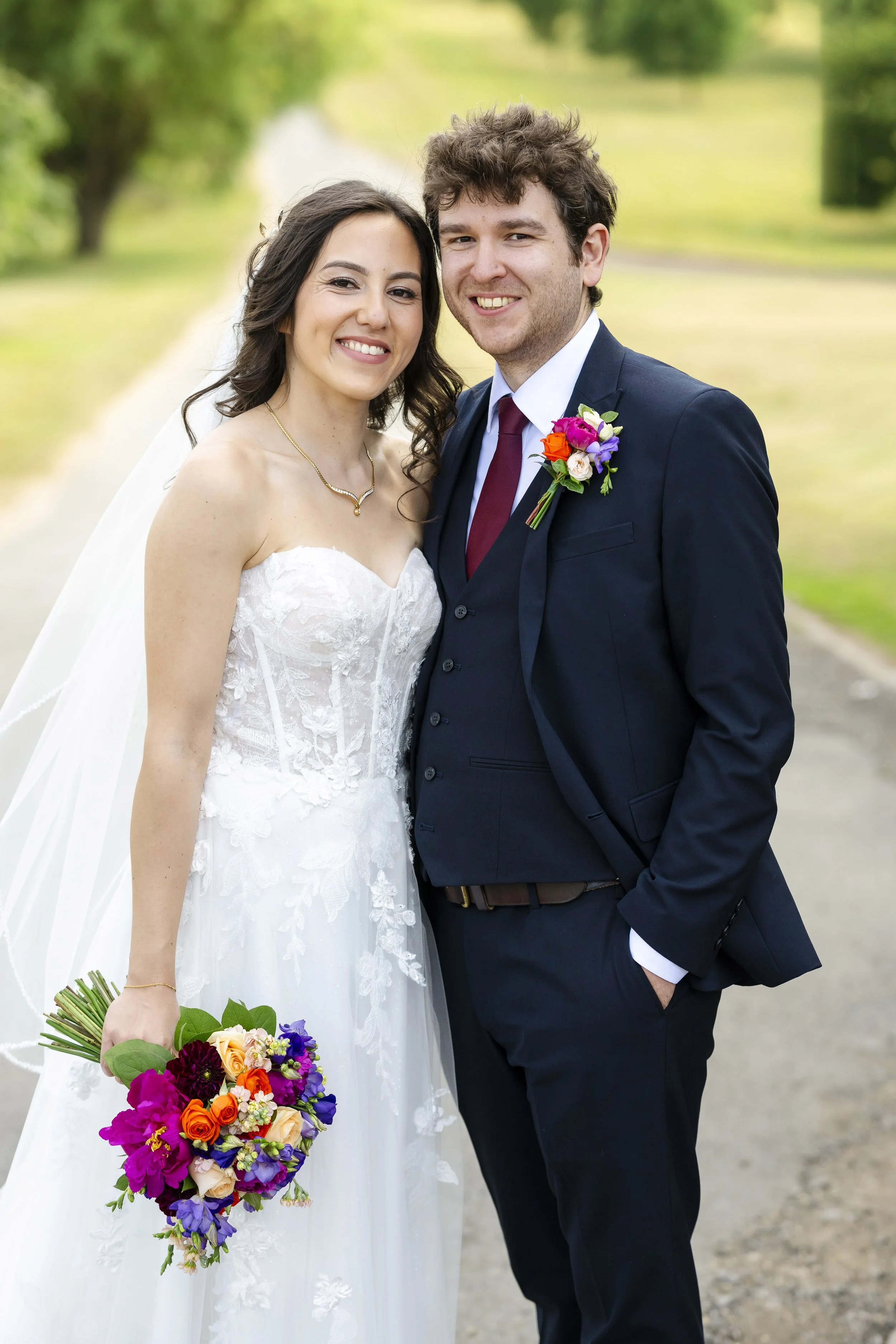 A newlywed couple smiling outdoors; the bride wears a strapless white wedding gown, holds a colorful bouquet, and has dark, curly hair; the groom wears a navy suit, white shirt, burgundy tie, and has a boutonniere; background features greenery and a 