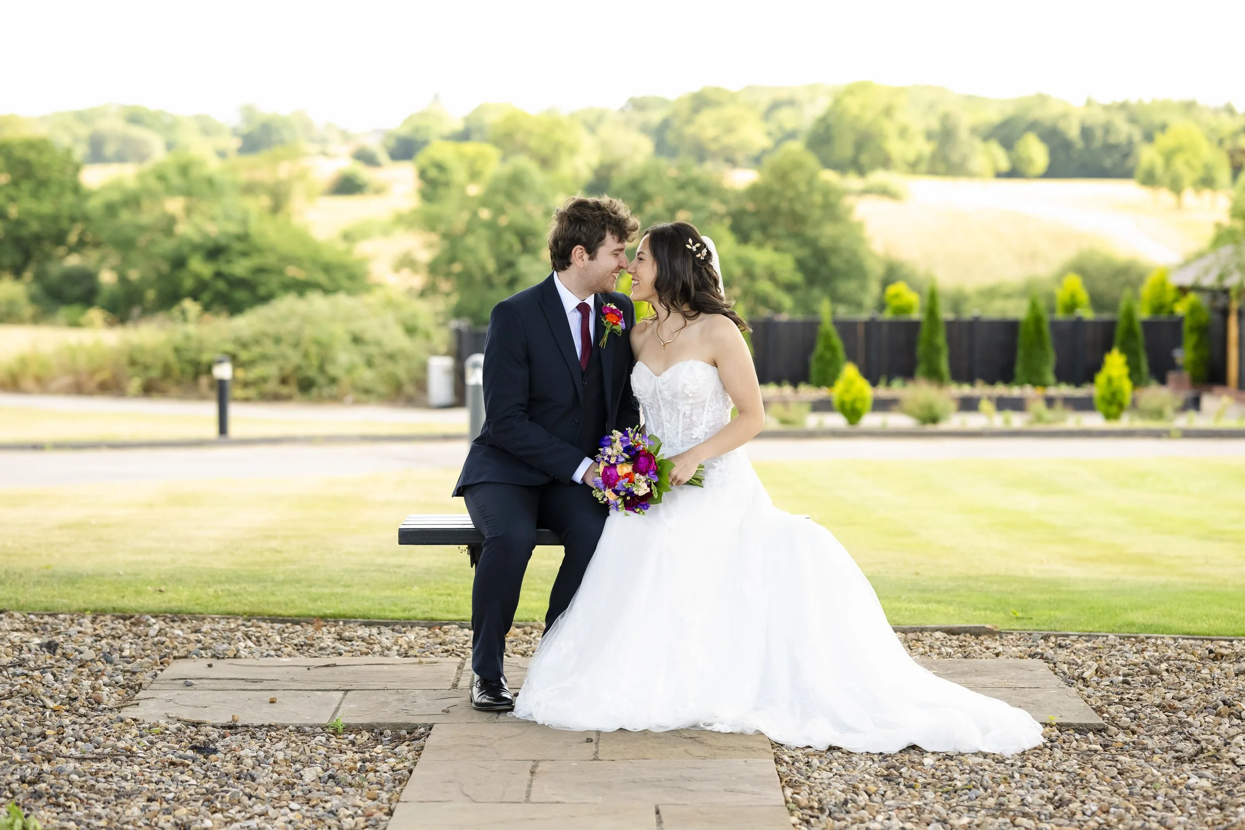 A bride and groom sitting on a park bench, leaning in with foreheads touching, holding a colorful bouquet, outdoors on a sunny day, with green trees and rolling hills in the background.