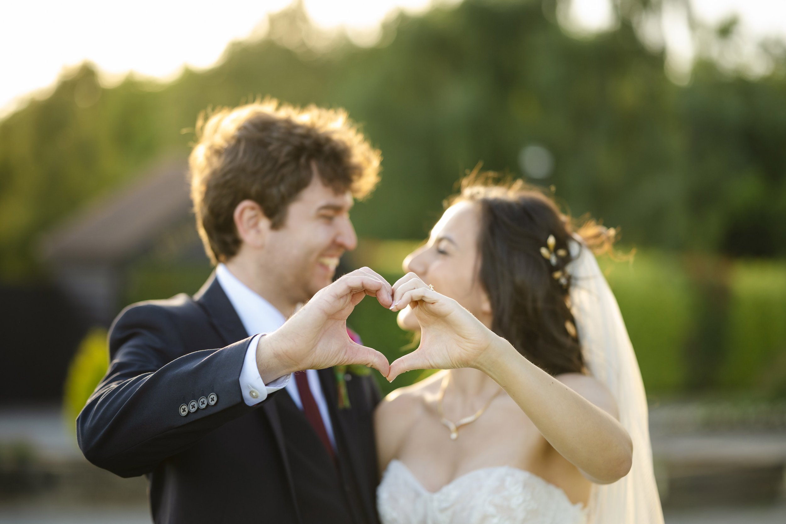 A bride and groom smiling and making a heart shape with their hands on their wedding day outdoors during sunset.