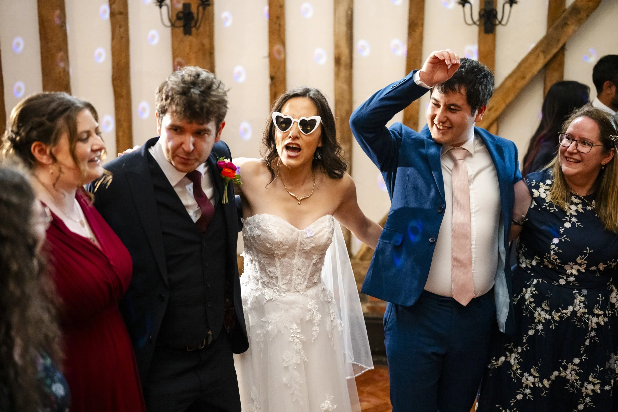 People celebrating at a wedding reception, the bride wearing a white wedding dress and sunglasses, the groom in a blue suit and pink tie, and friends standing in a circle, smiling and dancing.