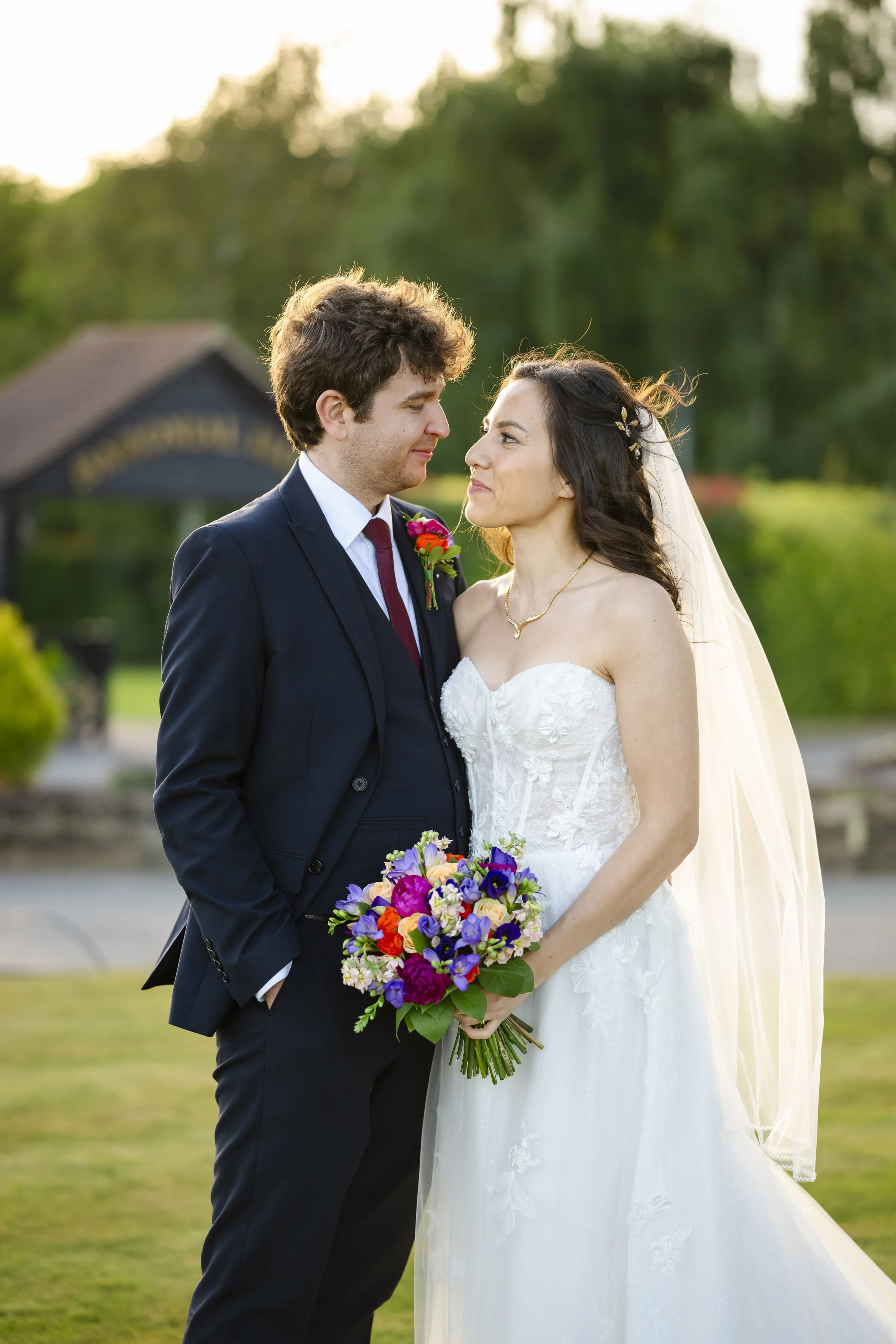 A bride and groom stand close together outdoors during sunset, looking at each other with a background of greenery and a small building.