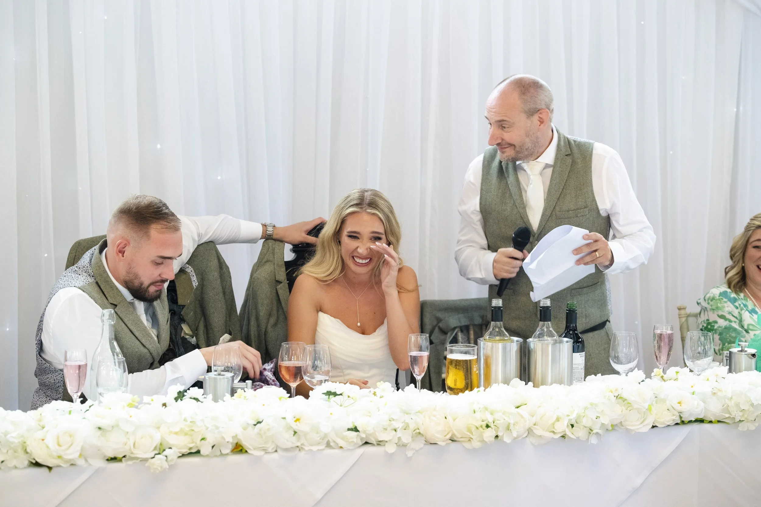 A woman in a wedding dress laughing while seated at a decorated wedding table with two men, one giving a speech, and other guests in the background.
