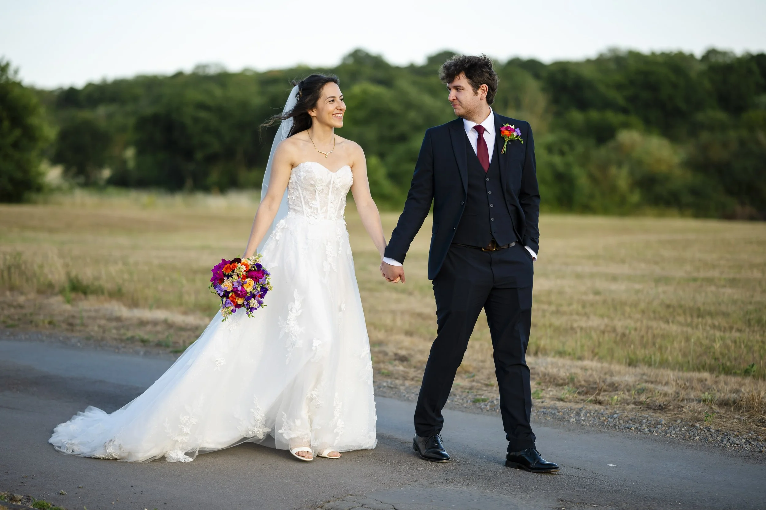 A bride and groom holding hands while walking outdoors on their wedding day, with greenery in the background.