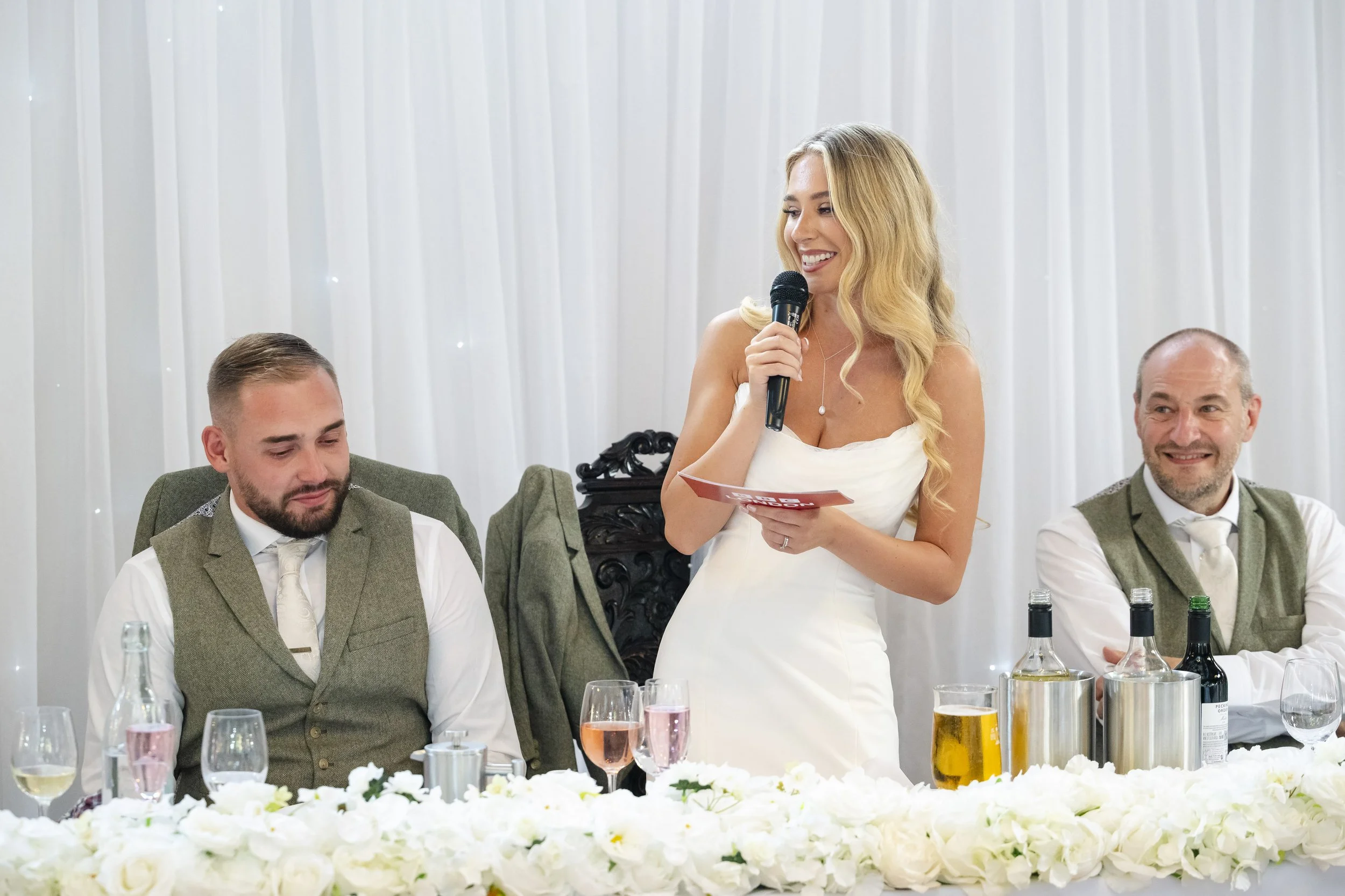 A woman in a white dress giving a speech at a wedding reception, standing beside two seated men in suits, with drinks and flowers on the table.