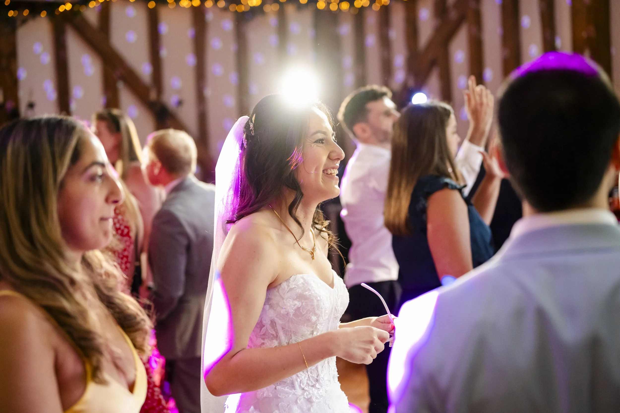 A bride at her wedding reception, smiling and holding a straw, surrounded by guests under colorful party lights.