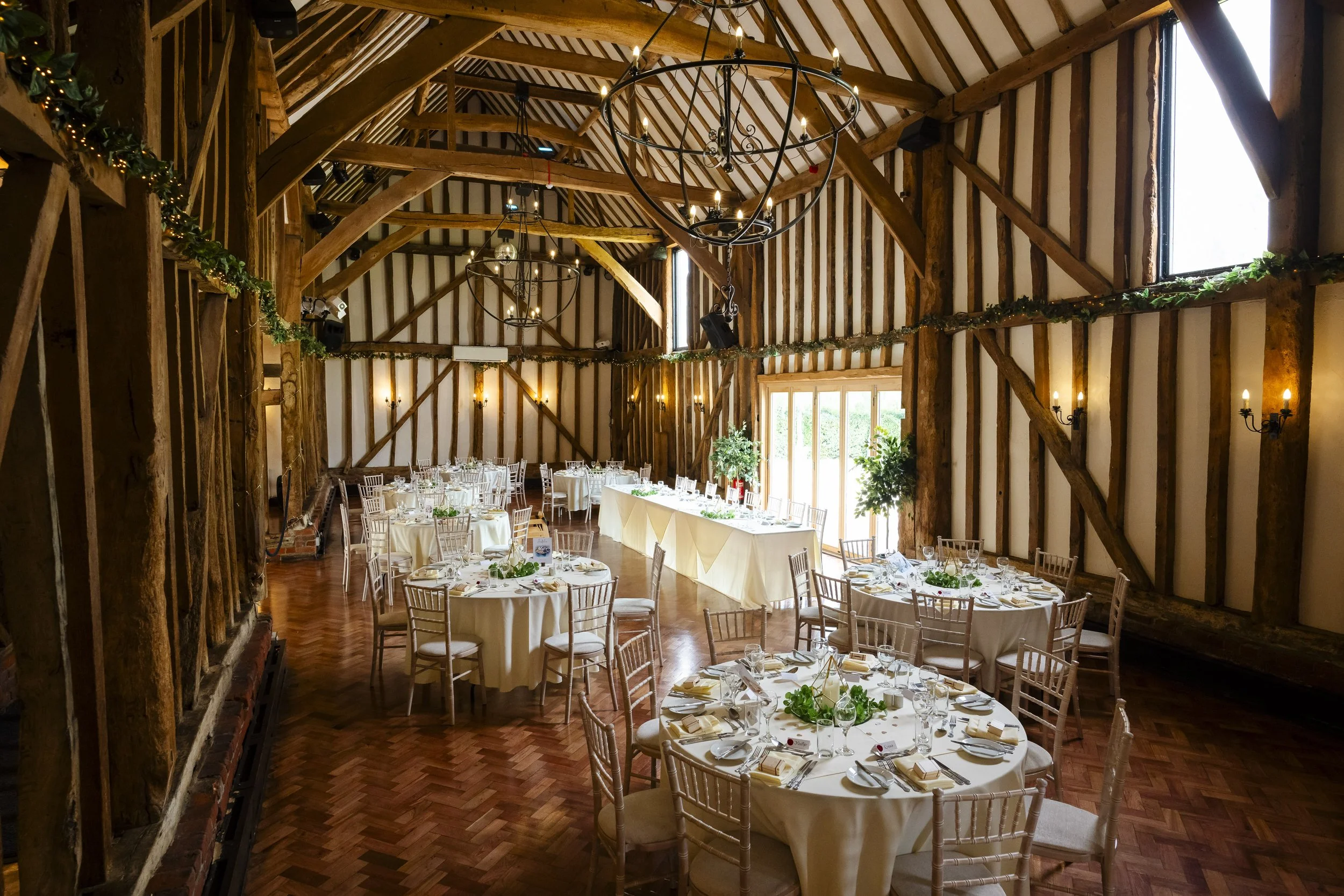 Elegant event space in a rustic barn with round dining tables, white tablecloths, and gold chairs, decorated with greenery, chandeliers, and wall sconces, set for a formal dinner.