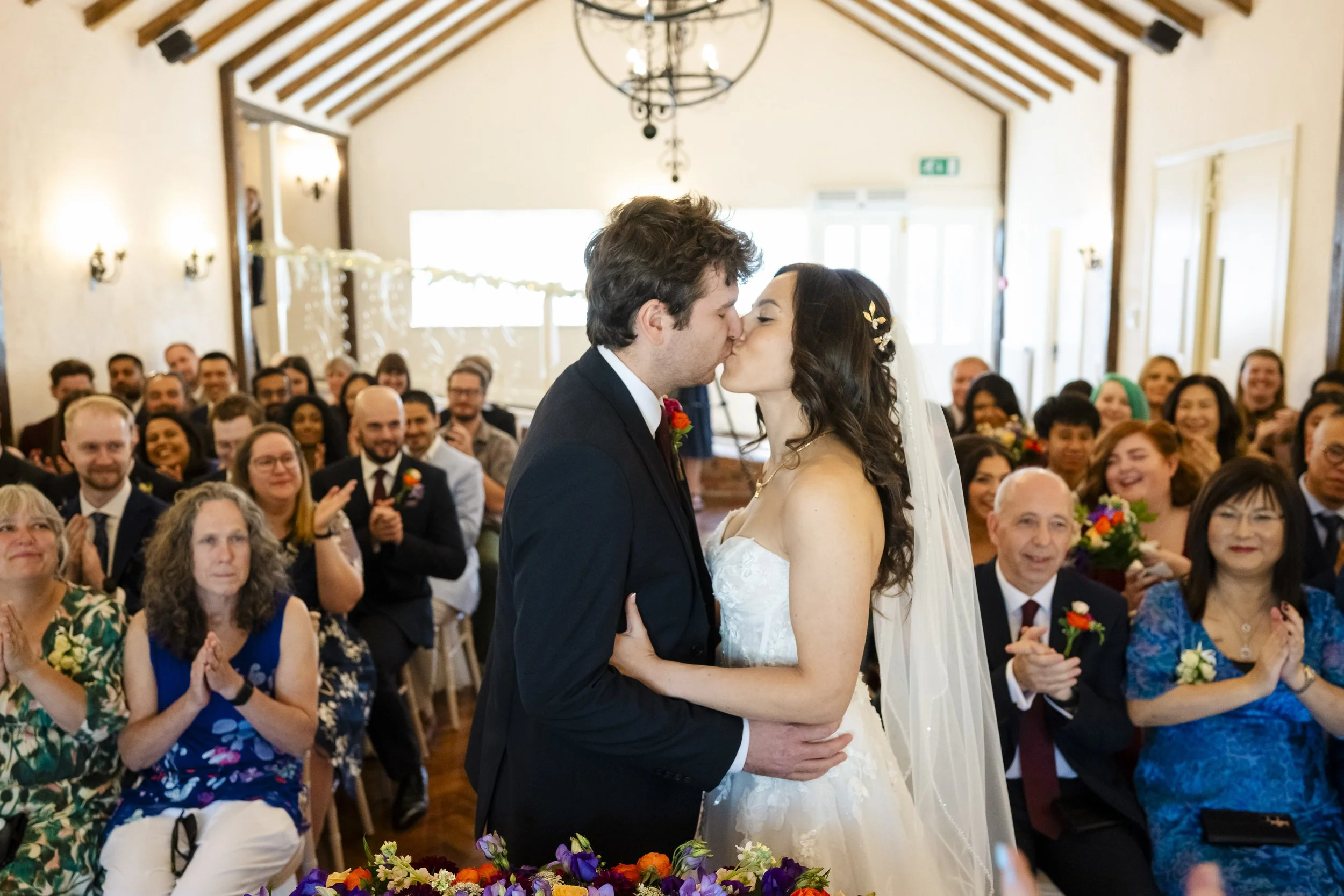 A bride and groom kiss during their wedding ceremony, surrounded by smiling guests in an indoor venue with wooden ceiling beams.