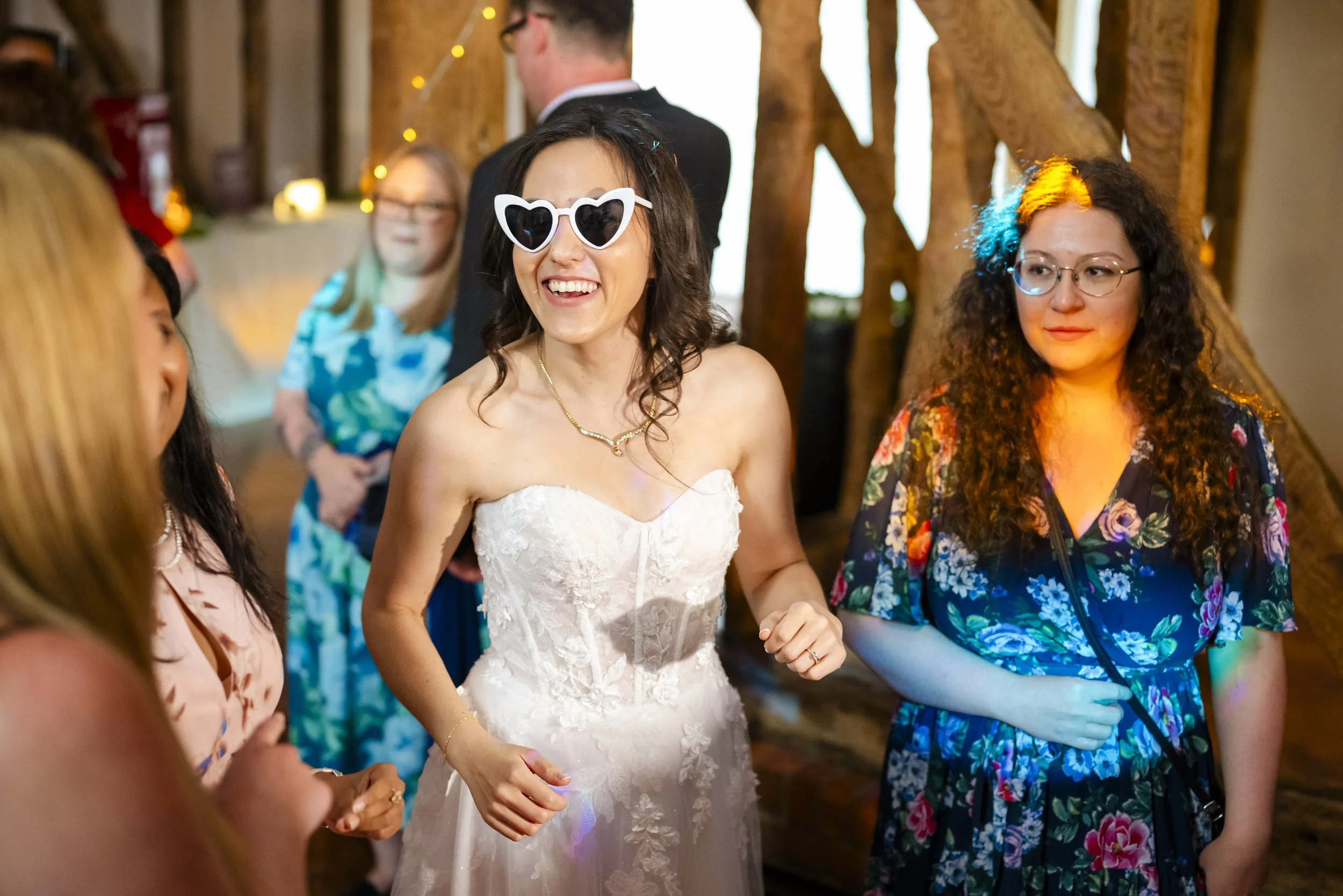 A woman in a wedding dress wearing heart-shaped sunglasses is smiling and talking with others at a celebration. The setting appears to be a rustic indoor venue with wooden beams and warm lighting.