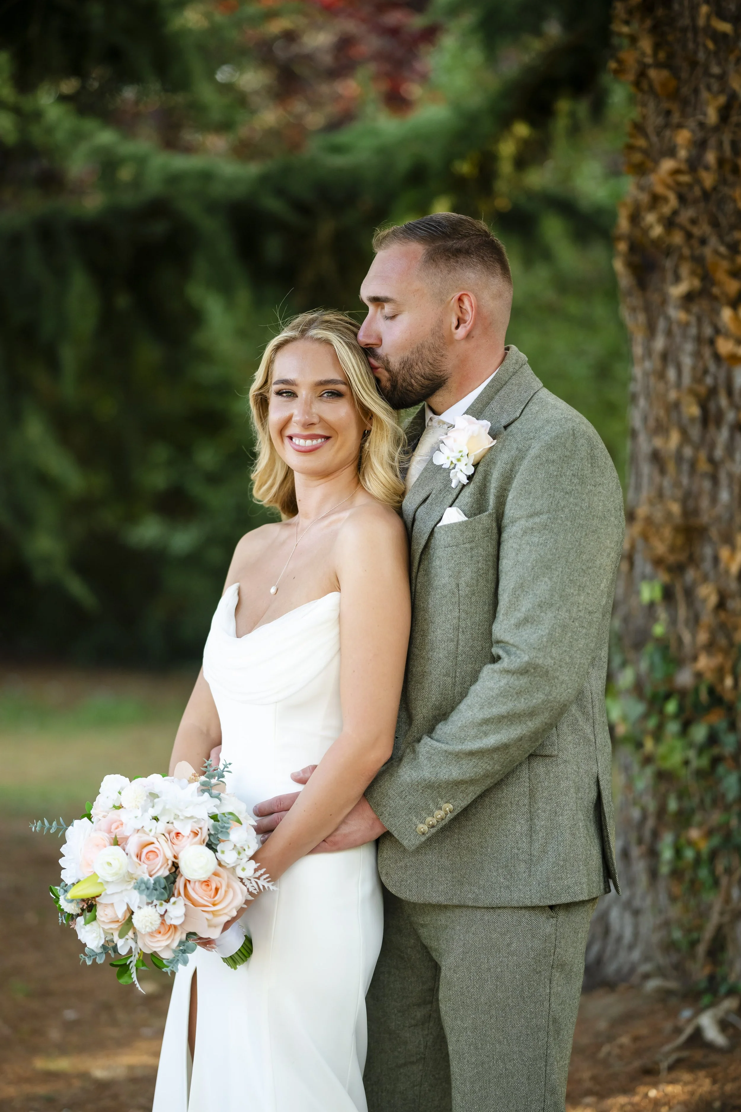 A bride and groom standing outdoors by a large tree, smiling and sharing a tender moment. The bride is wearing a strapless white wedding dress and holding a bouquet of white and peach roses. The groom is in a grey suit with a boutonniere. The backgro