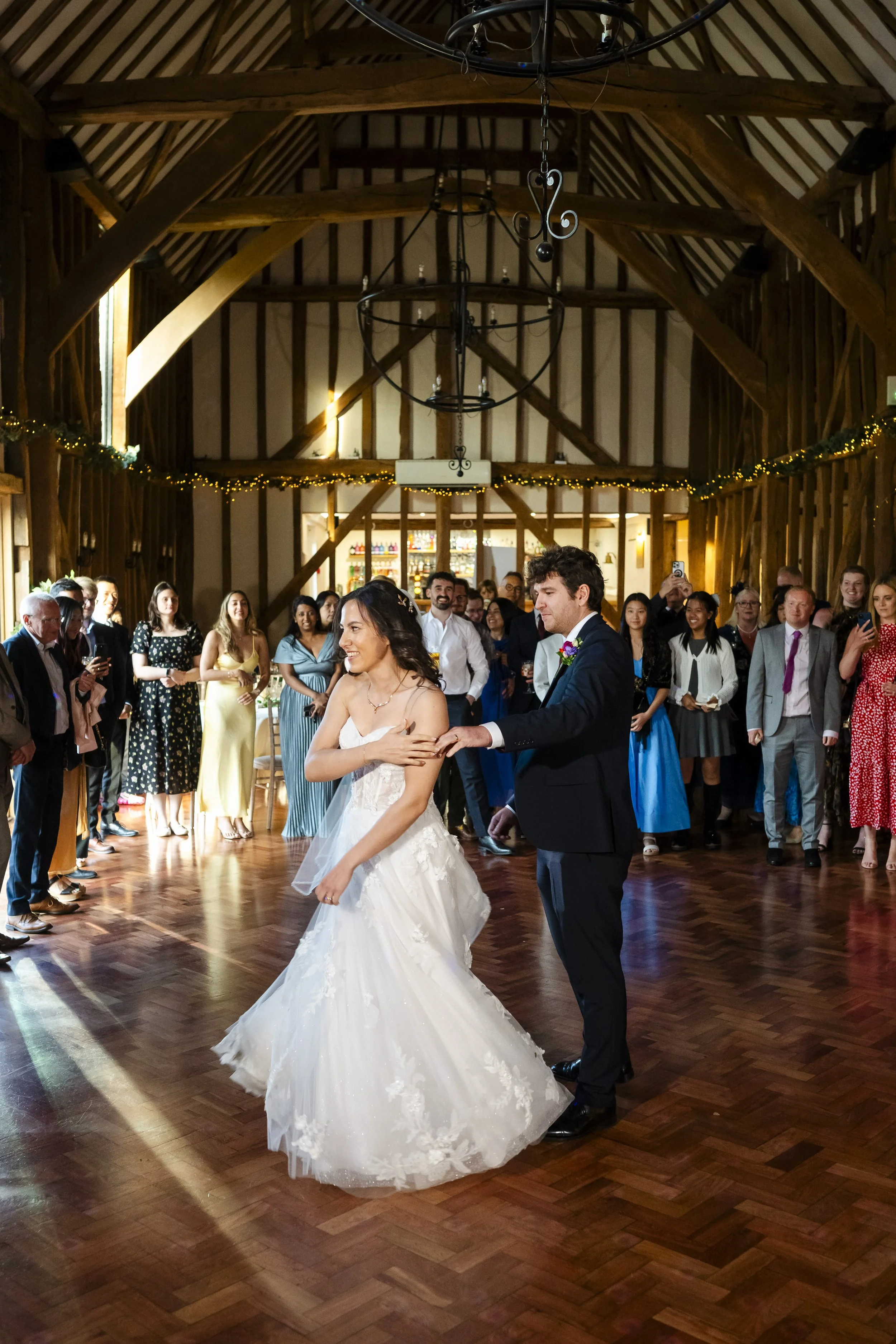 A bride and groom are dancing at their wedding reception in a rustic barn with wooden beams. Guests are watching and taking photos.