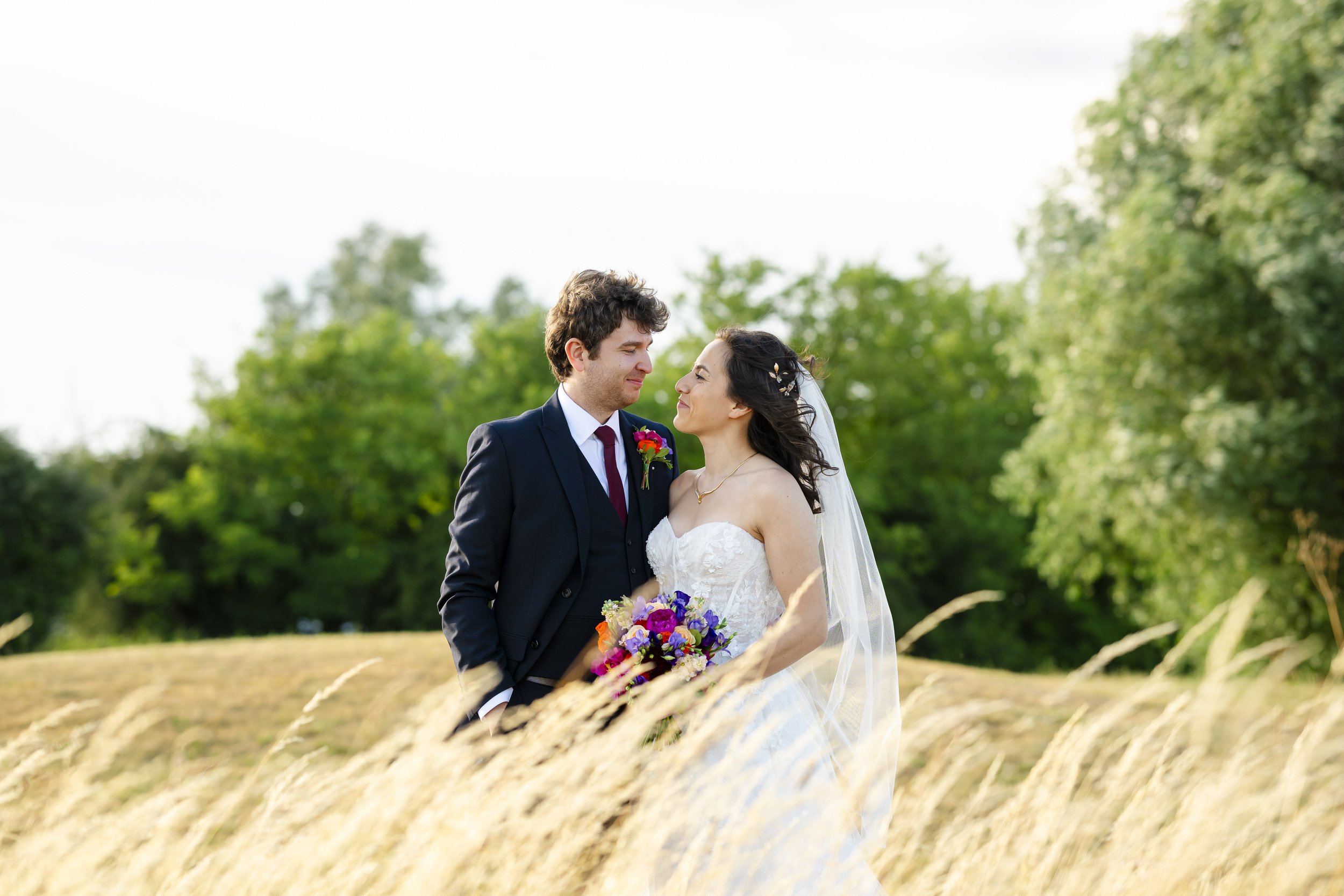 A bride and groom standing close outdoors in a field with tall grass, facing each other lovingly, with trees and a bright sky in the background.