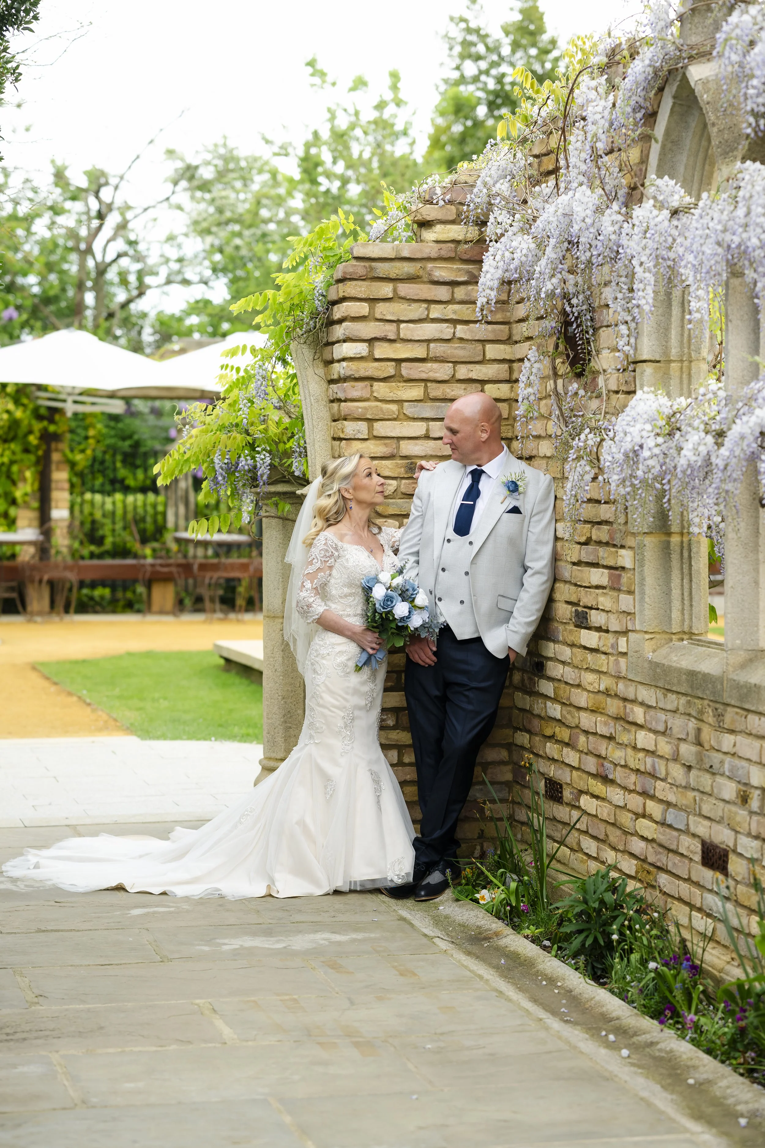 A bride and groom standing close together by a brick wall with purple wisteria flowers. The bride wears a white lace wedding dress and holds a bouquet, the groom wears a light-colored suit jacket, dark pants, and a dark tie, looking at each other in 