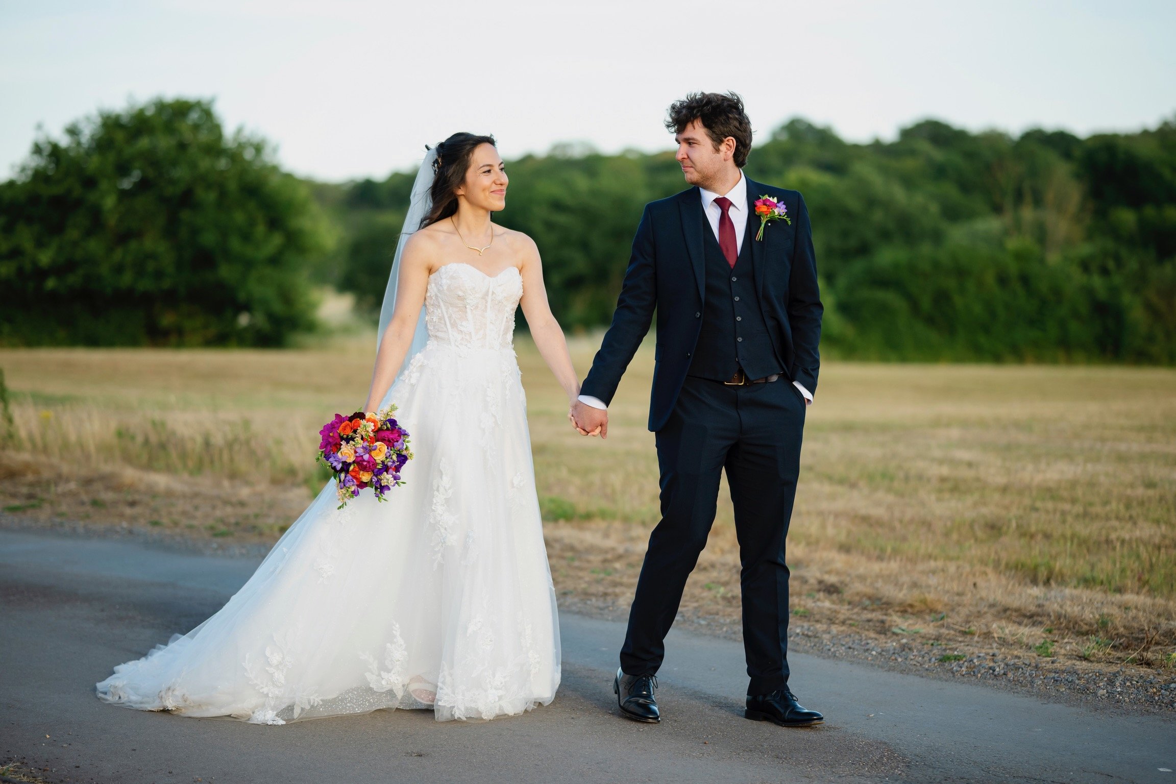 A bride and groom holding hands and walking outdoors, with the bride in a wedding dress holding a colorful bouquet and the groom in a dark suit with a boutonniere, during daylight.