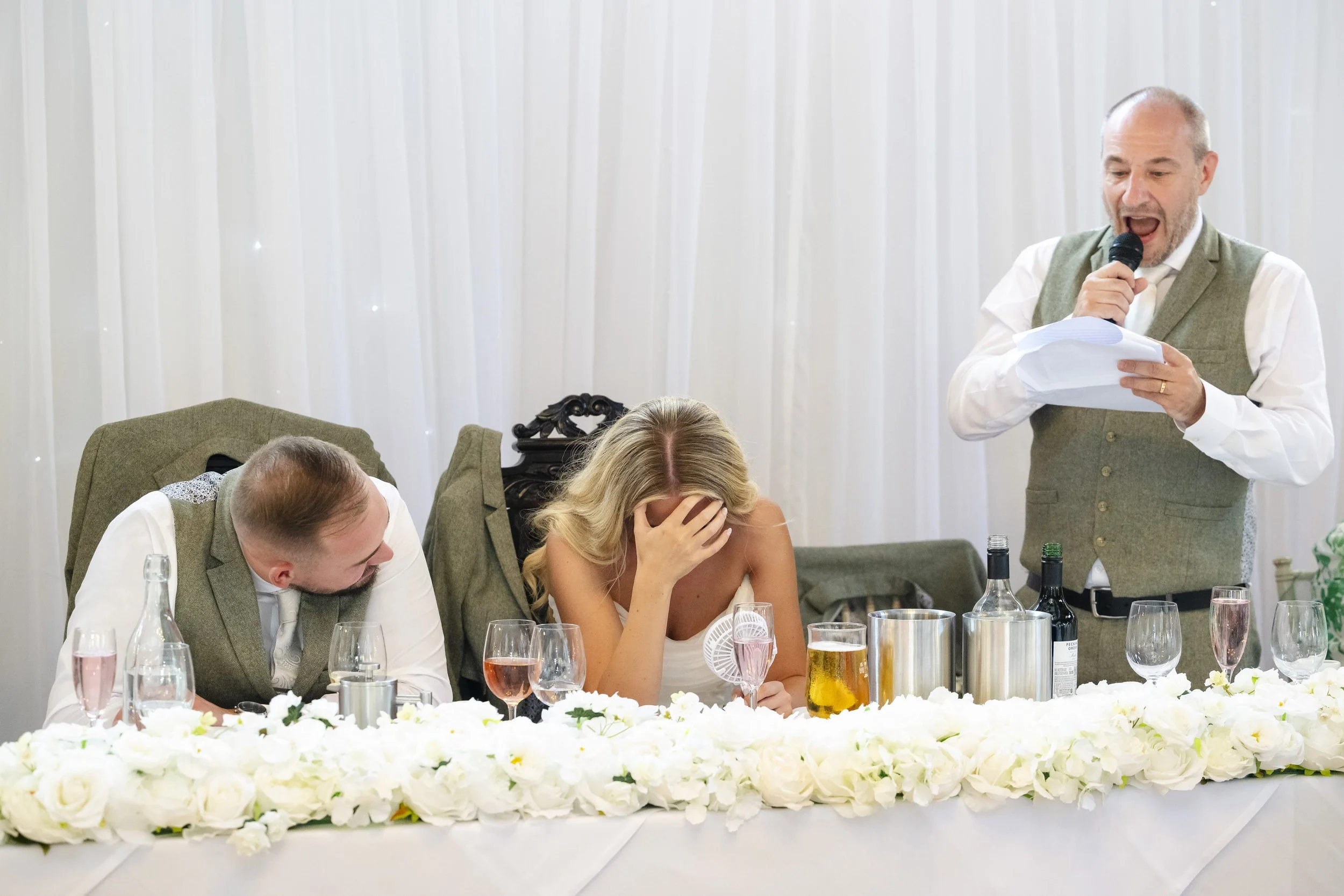 A bride in a white wedding dress sitting at a table with her head in her hand, appearing emotional. A groom sits next to her, leaning toward her, both at a wedding reception. An older man stands, speaking into a microphone and reading from a paper.