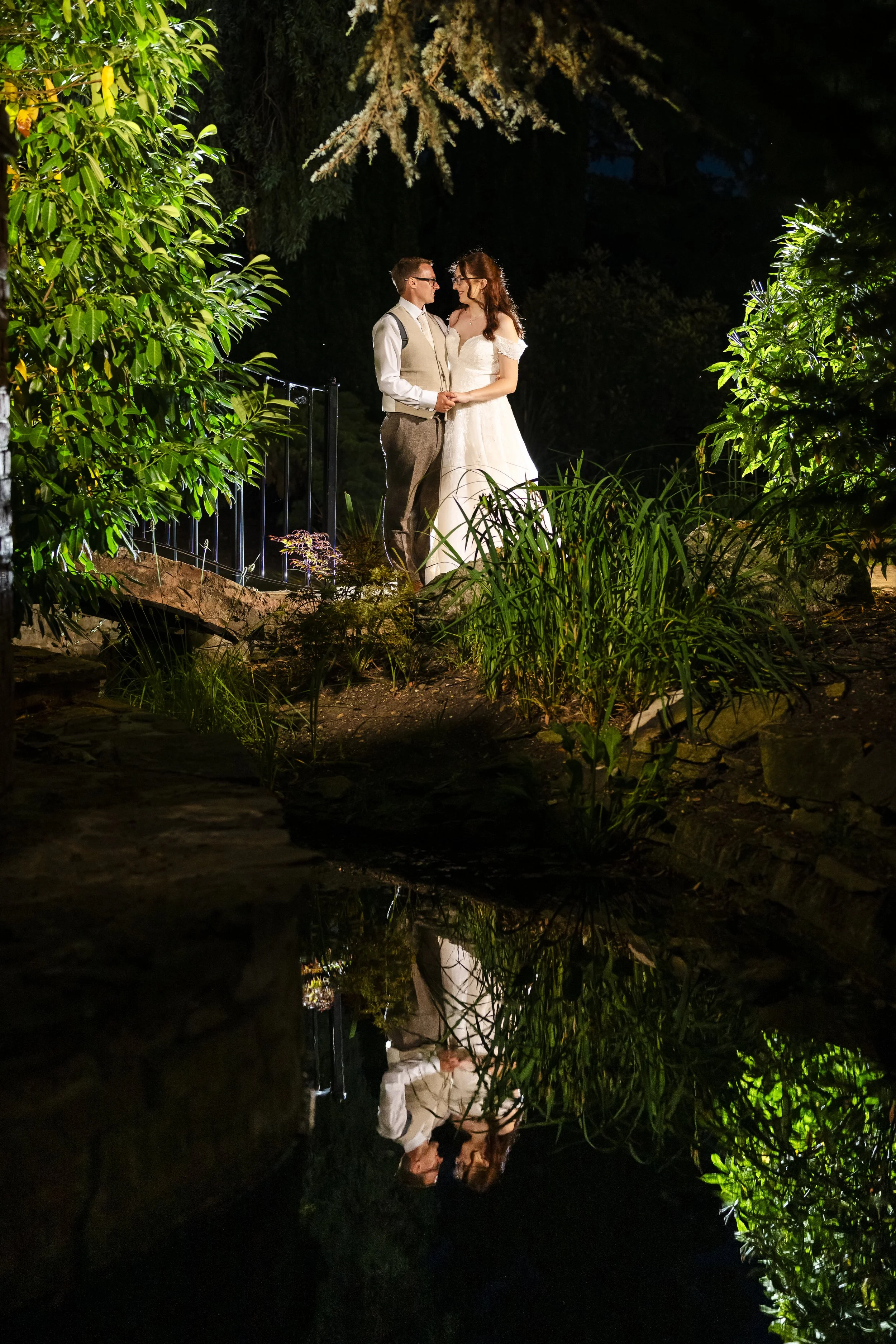 A bride and groom holding hands and looking at each other at night in a lush, green outdoor setting with a small pond reflecting their image.