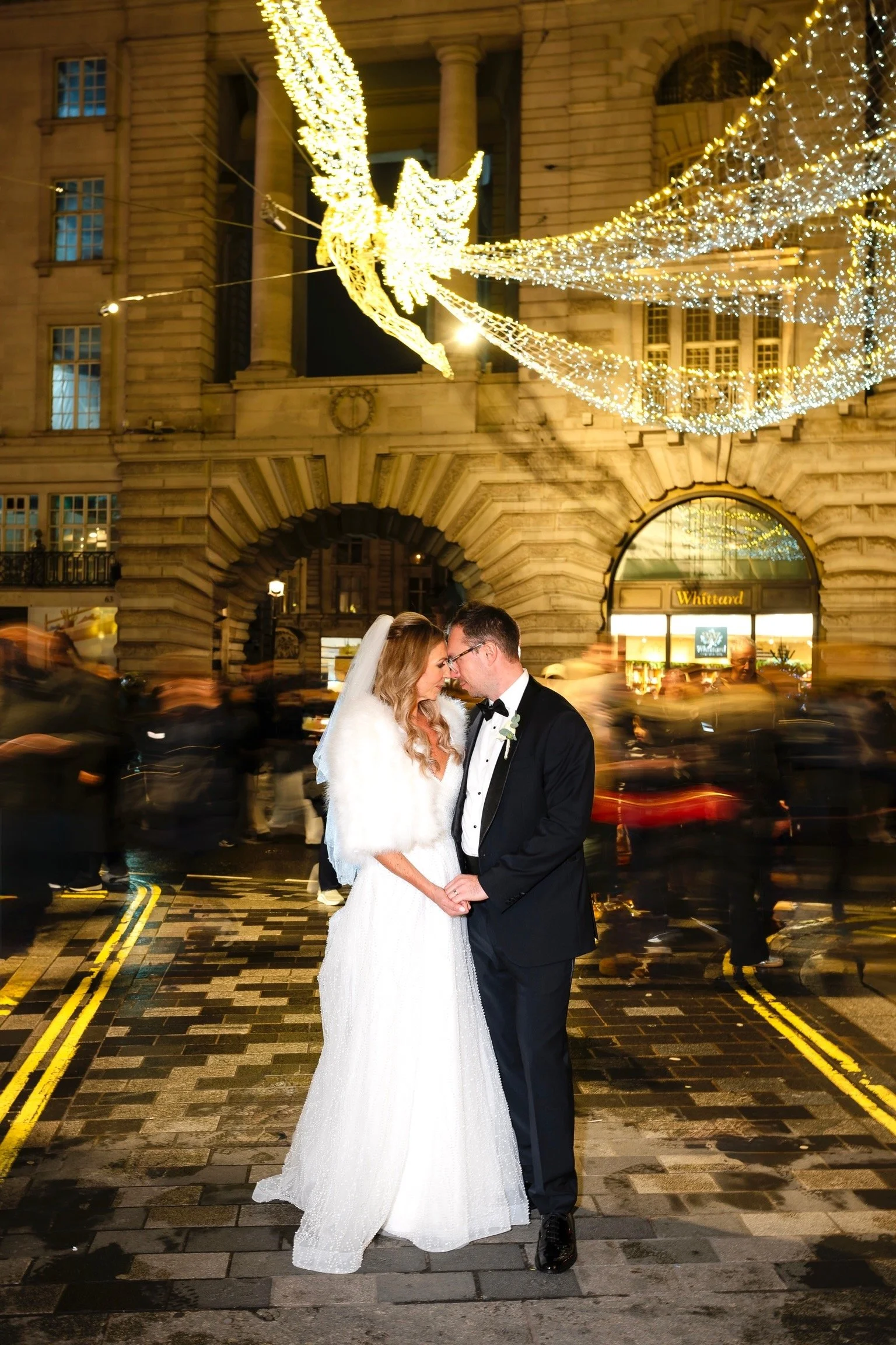 A bride and groom standing close together, touching foreheads, in front of a historic stone building decorated with festive Christmas lights, during what appears to be a wedding celebration at night.