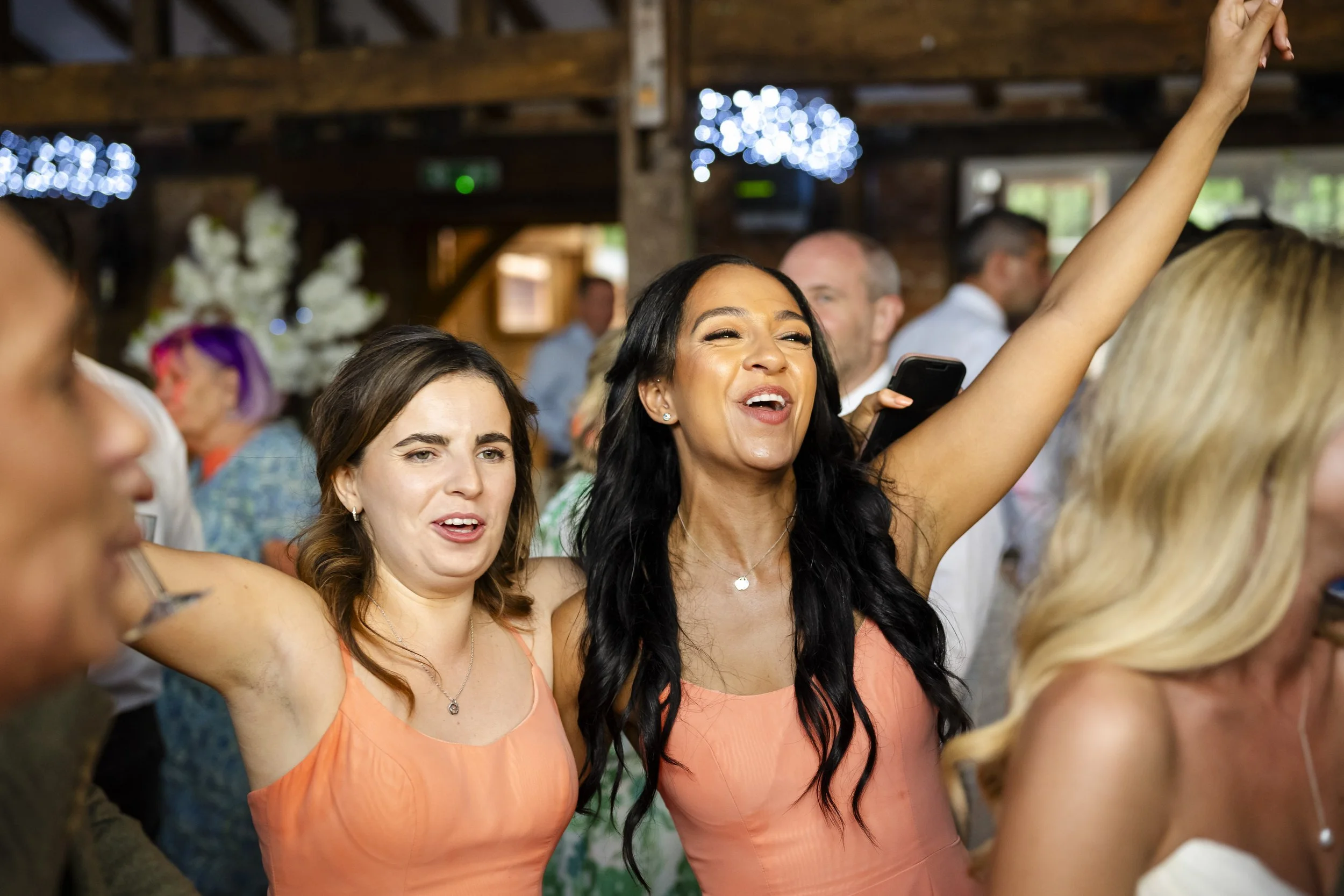 A woman with long black hair wearing a peach dress is smiling and raising her arm in celebration at a social event or party. She is surrounded by other guests, some of whom are also smiling and enjoying the moment.