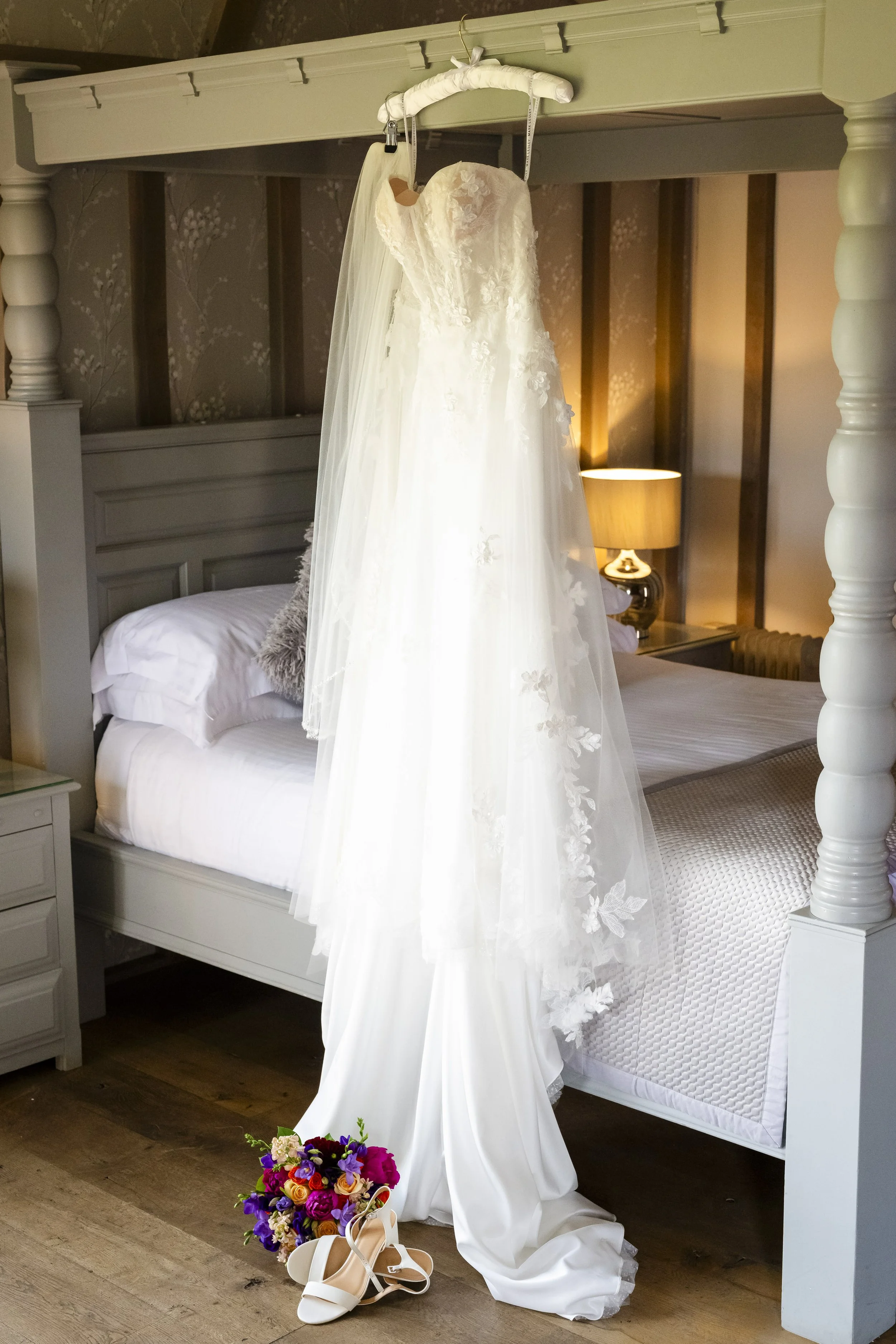 A wedding dress with a veil hangs from a canopy above a bed. On the floor, there are white high-heeled shoes next to a bouquet of colorful flowers.