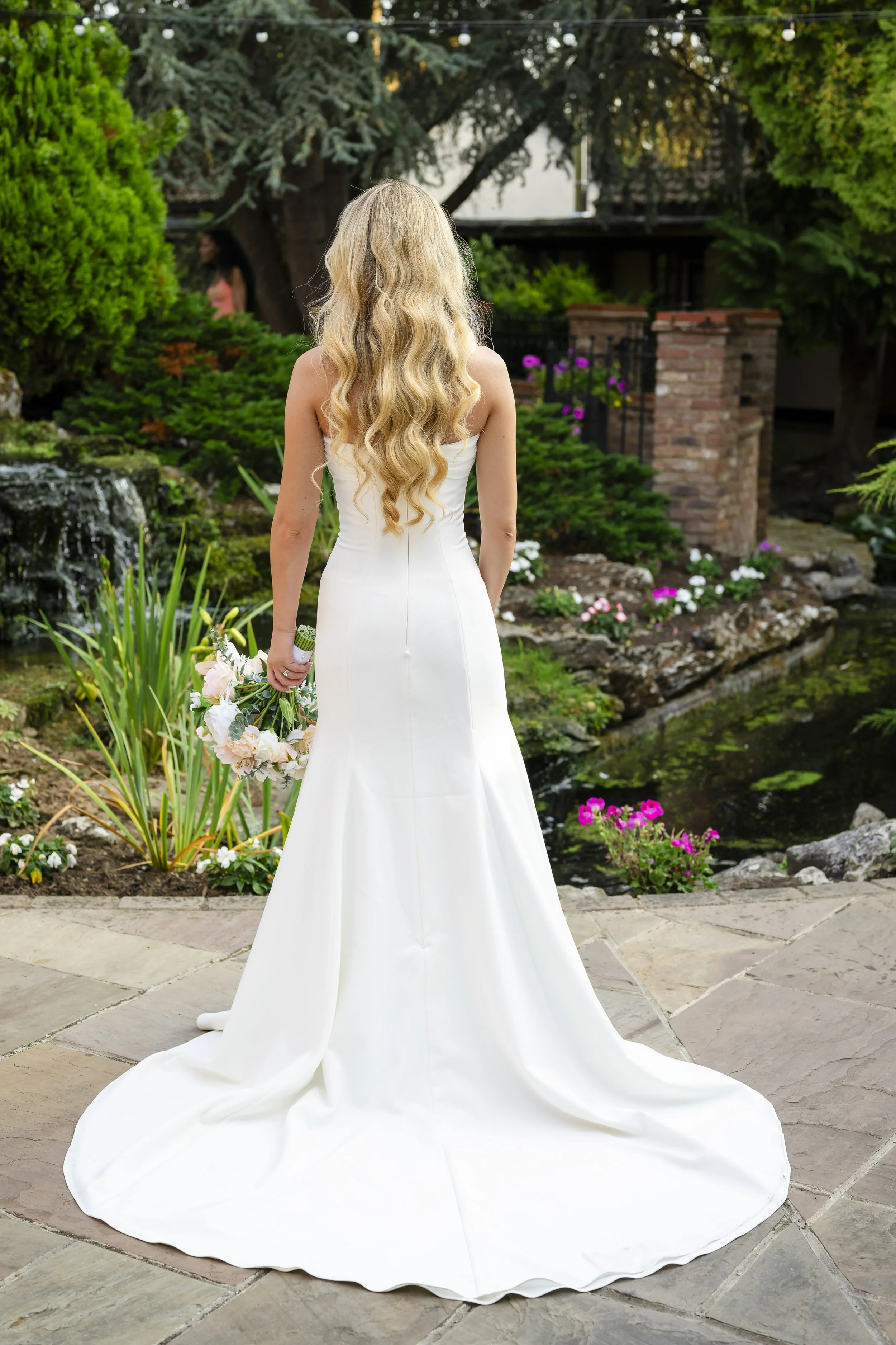 A bride in a white wedding gown holding a bouquet, standing outdoors in a garden with flowers and greenery.