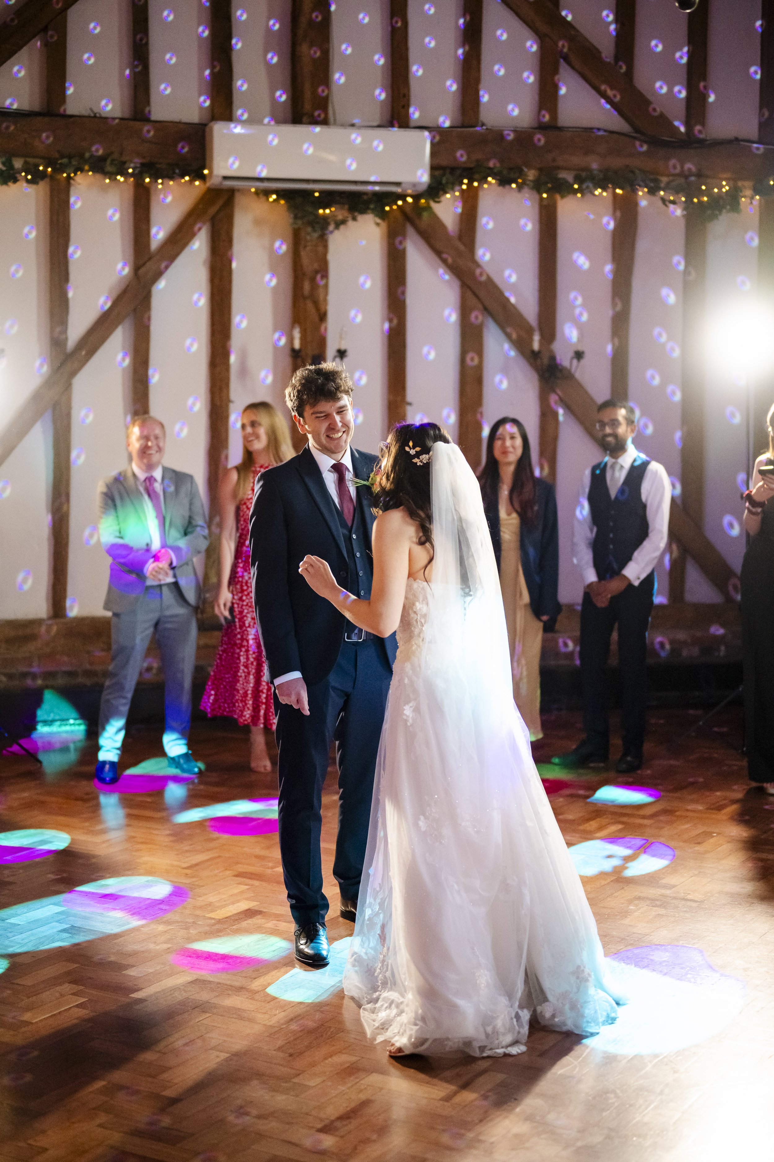 A bride and groom dancing during their wedding reception, with guests watching, colorful lights, and a decorated barn interior.