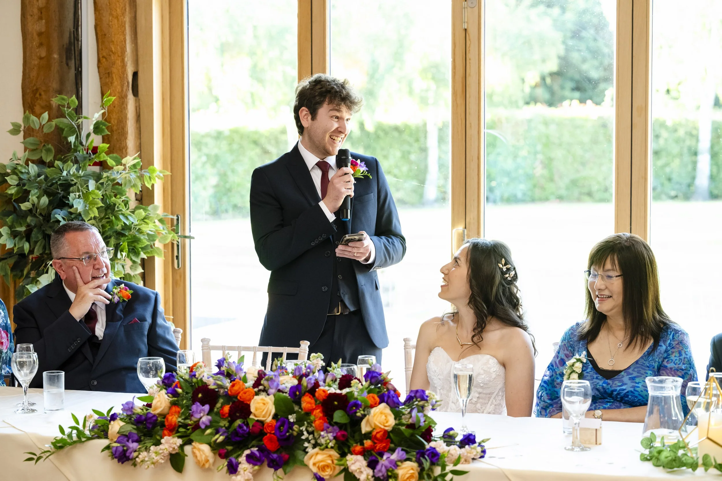A man in a black suit giving a speech at a wedding reception, with a bride and two women seated at the table, decorated with a large floral centerpiece.