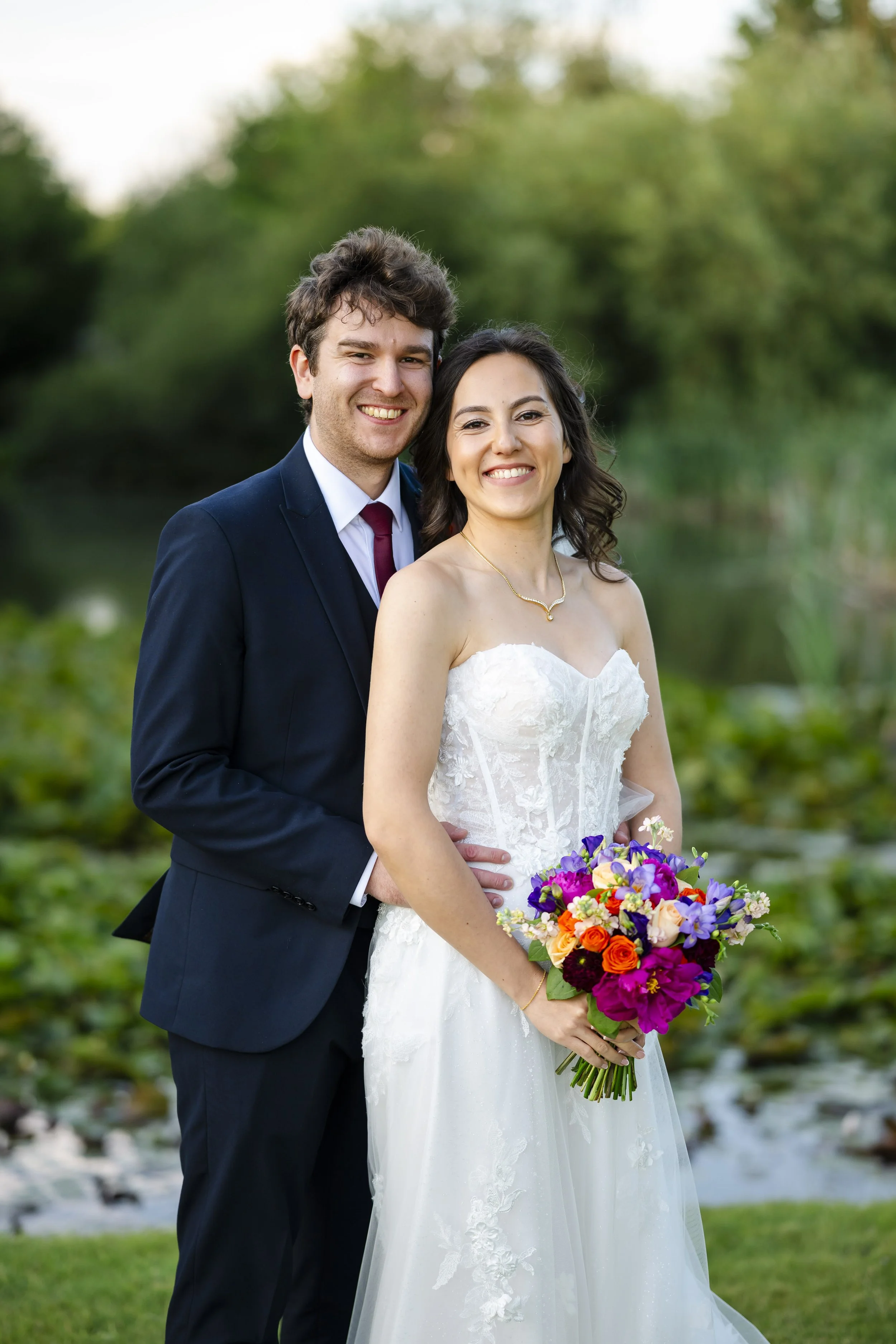A newlywed couple, a man in a navy suit and tie and a woman in a white strapless wedding gown, standing outdoors with greenery in the background. The woman is holding a colorful bouquet of flowers.