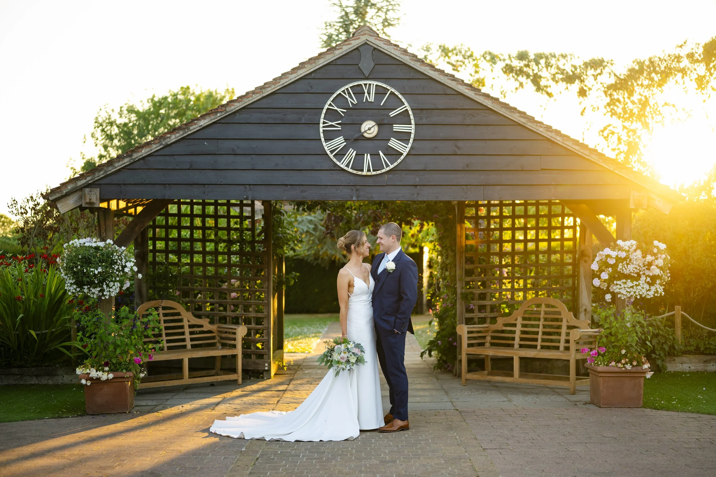 A wedding couple standing close together, facing each other, in front of a wooden garden archway with a clock on top, surrounded by colorful flowers and benches, during sunset.
