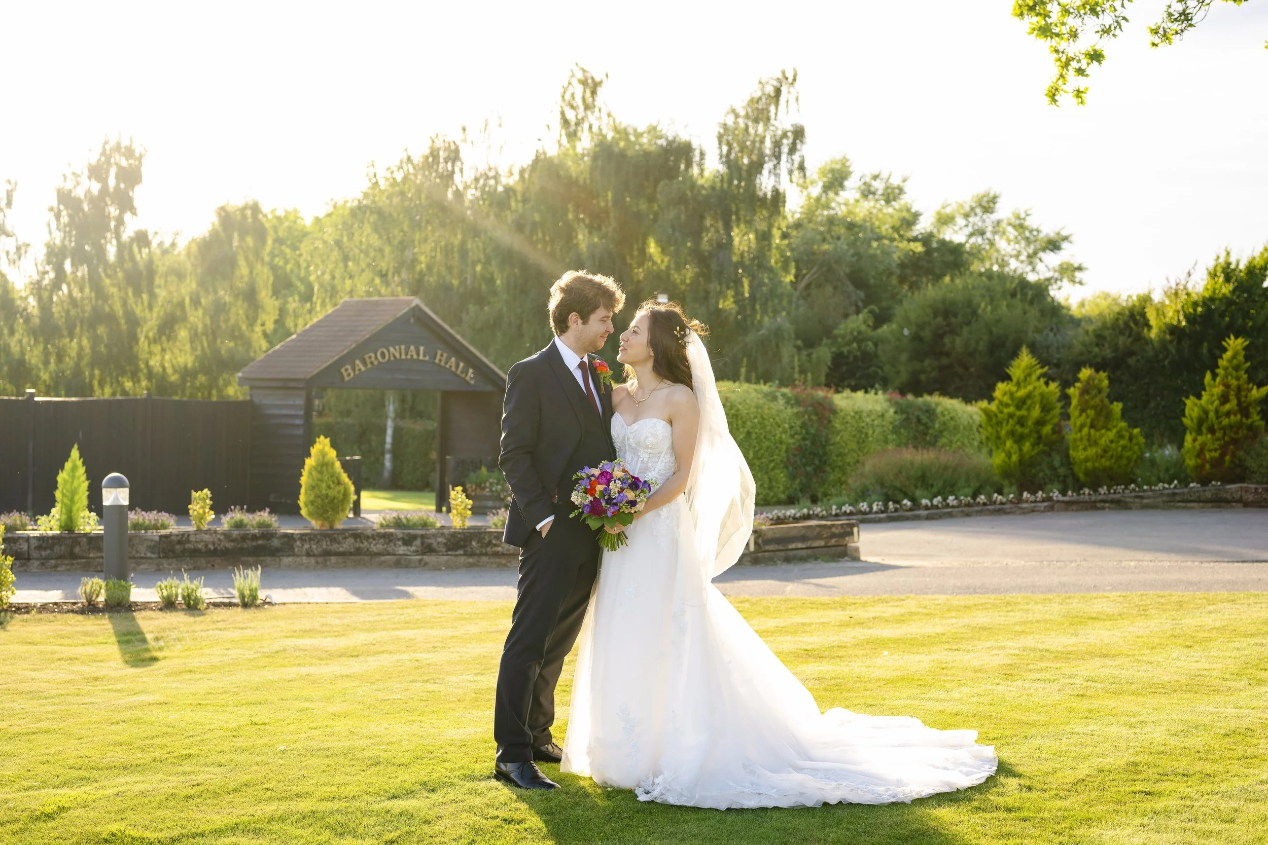 A bride and groom standing on a lawn, holding a bouquet, with the groom in a suit and the bride in a white wedding dress, during sunset at Baronal Hall.