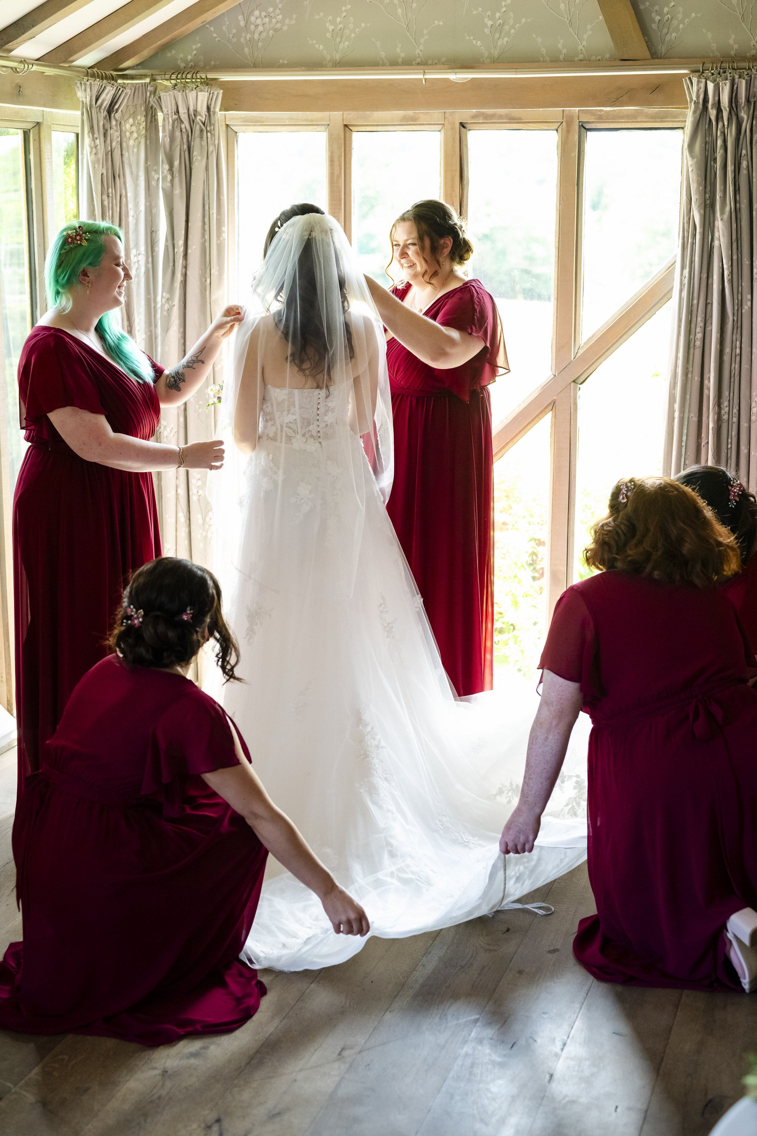 Bride in a white wedding dress is being assisted by her bridesmaids in a room with large windows and cream curtains, as they prepare for her wedding.