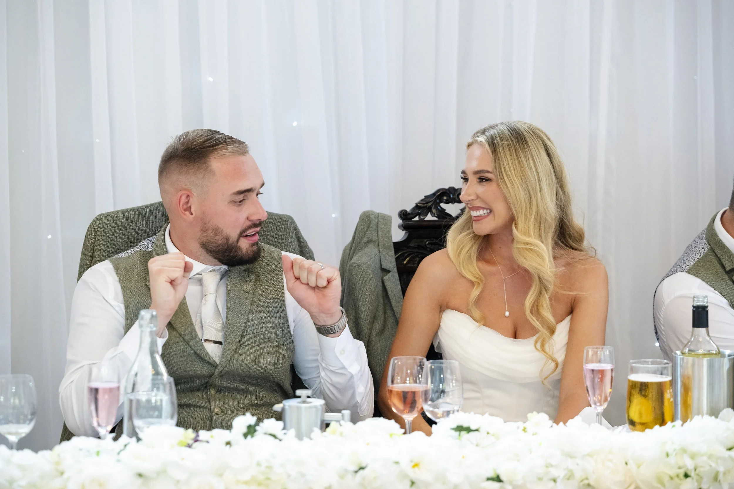 A bride and groom sitting at a wedding reception table, smiling and talking to each other, with glasses and bottles on the table and white drapery in the background.
