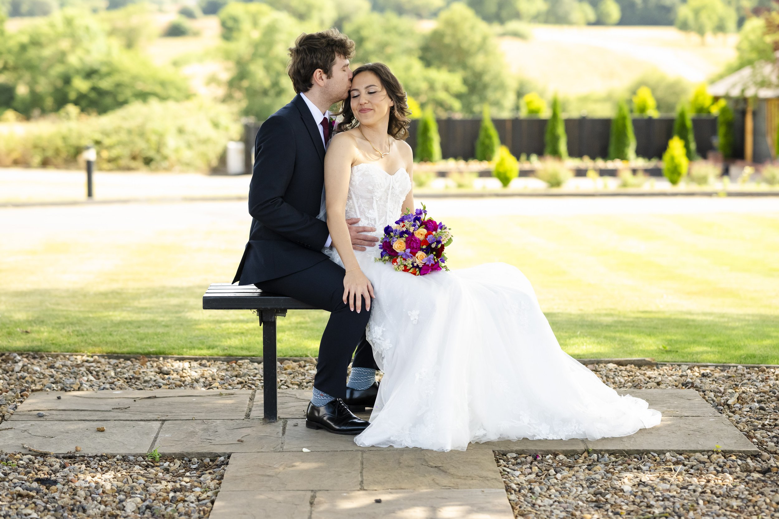 A bride and groom sit on a bench outdoors on a sunny day. The groom is dressed in a dark suit, and the bride is in a white wedding gown holding a colorful bouquet of flowers. The groom is kissing the bride's temple.