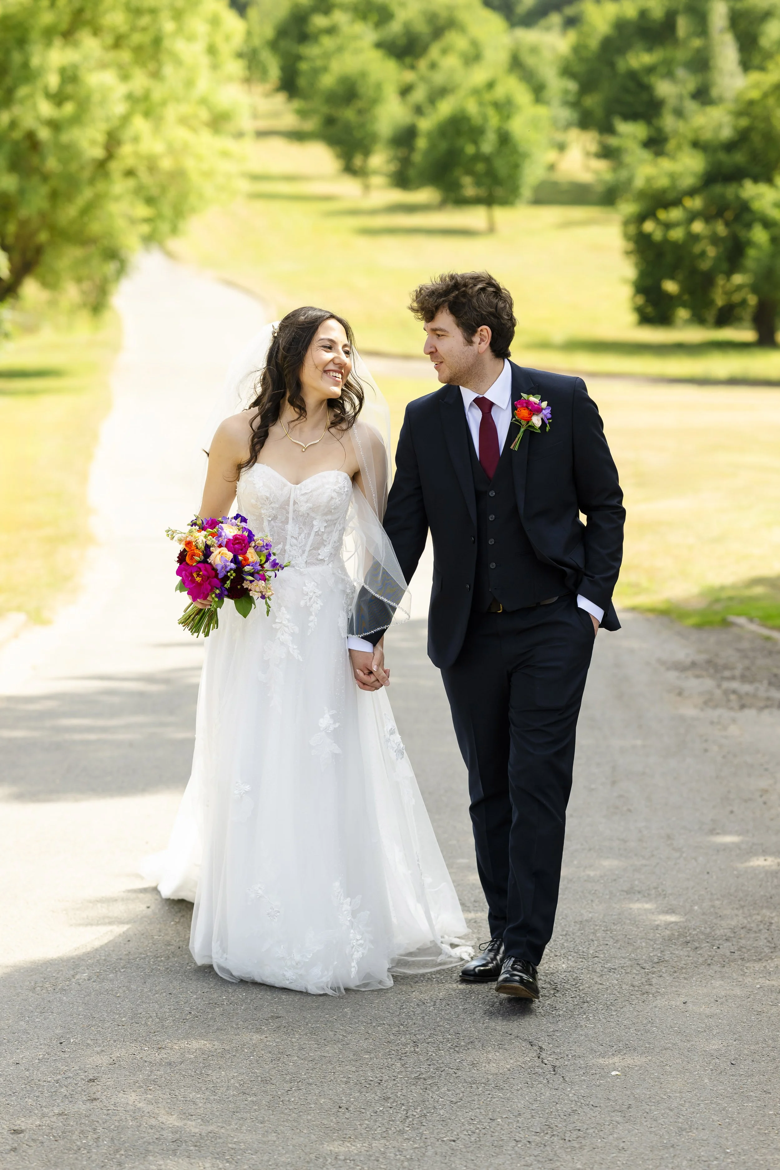 A bride and groom holding hands and walking together outdoors in a park with green trees and grass, celebrating their wedding day.