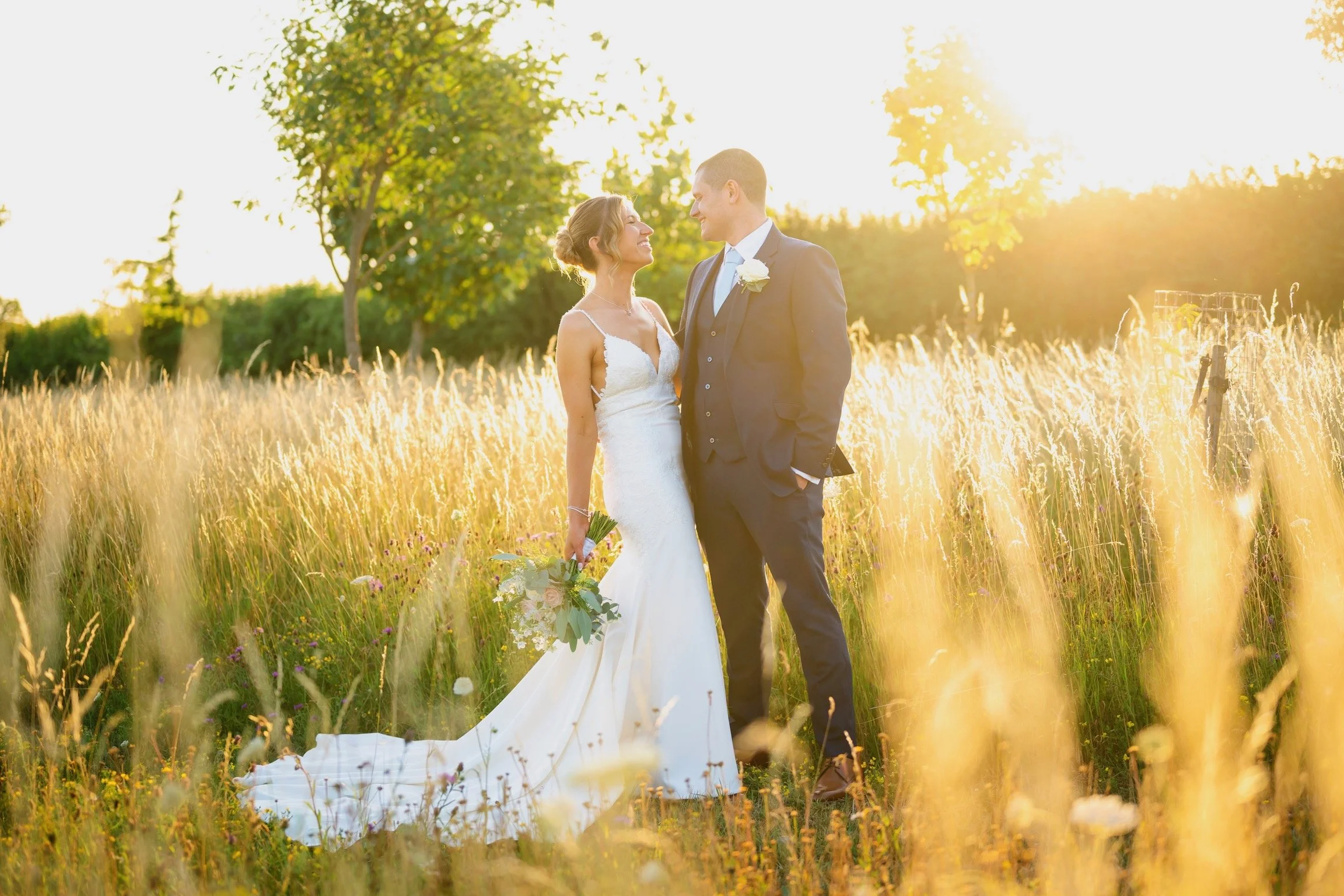 A bride and groom standing in a field of tall grass during sunset, gazing at each other while holding hands, with trees in the background.