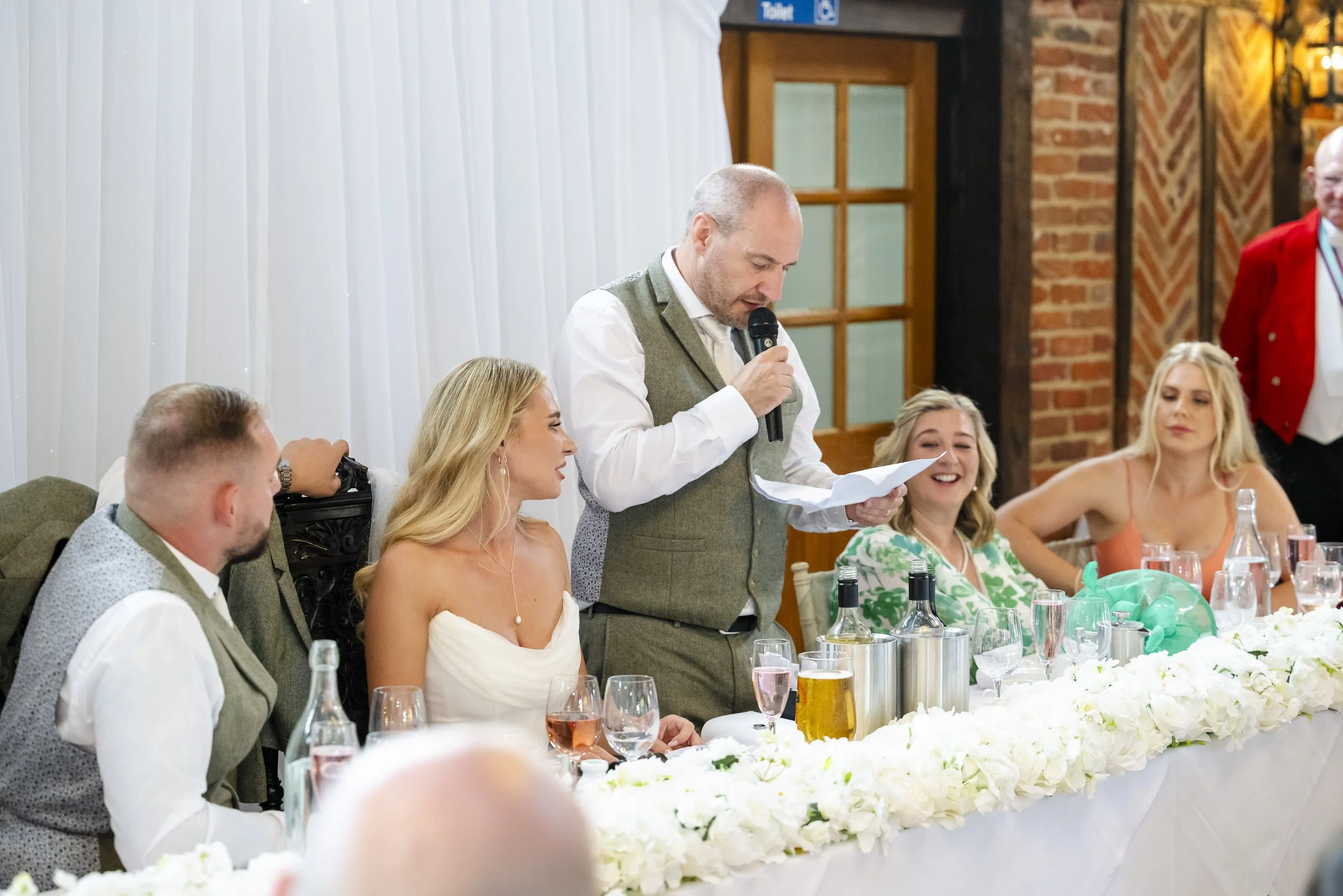 A man in a gray vest is giving a speech at a wedding reception. He is holding a microphone and reading from a paper. Seated at a decorated table with white flowers and drinks are several women and men, including a bride in a white strapless dress, an