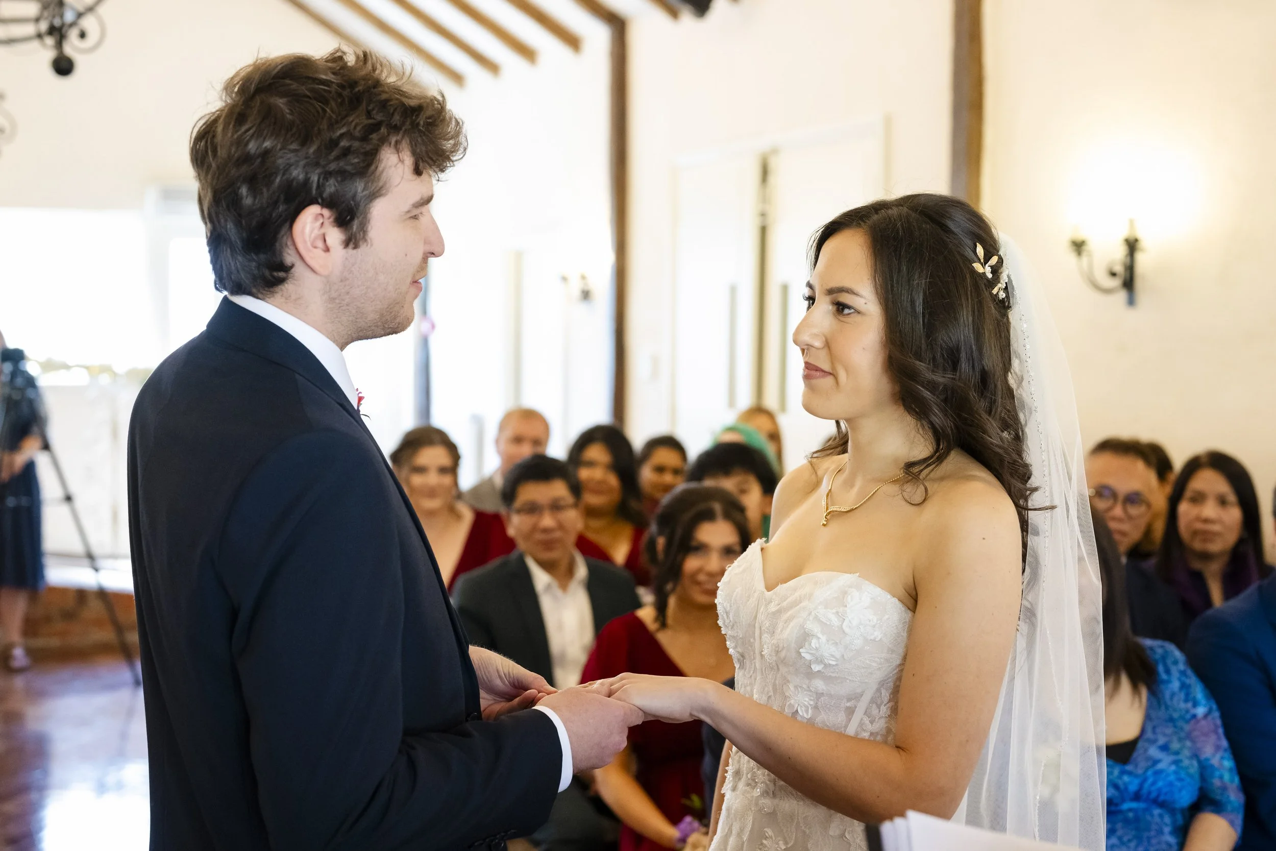 A bride and groom holding hands during their wedding ceremony, facing each other, with guests seated in the background.