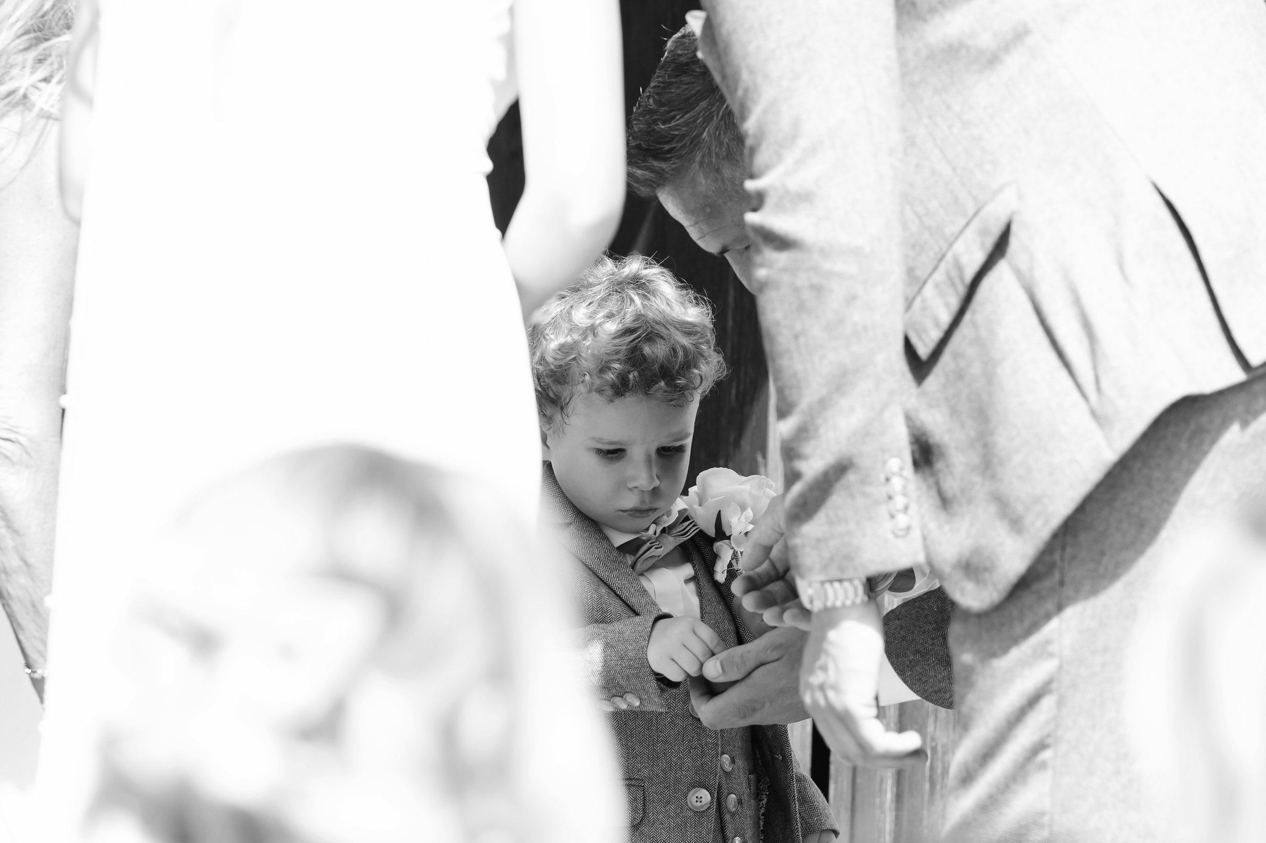 Young boy in a suit with a bow tie looking down at a boutonniere being pinned to his lapel during a wedding or formal event, with an adult assisting him.