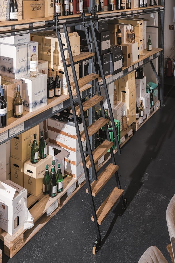 A store shelf filled with boxes of wine bottles and some bottles displayed outside the boxes. A wooden step ladder with black metal frame is placed in front of the shelves, which are made of wood and black metal support. The store has a dark floor.