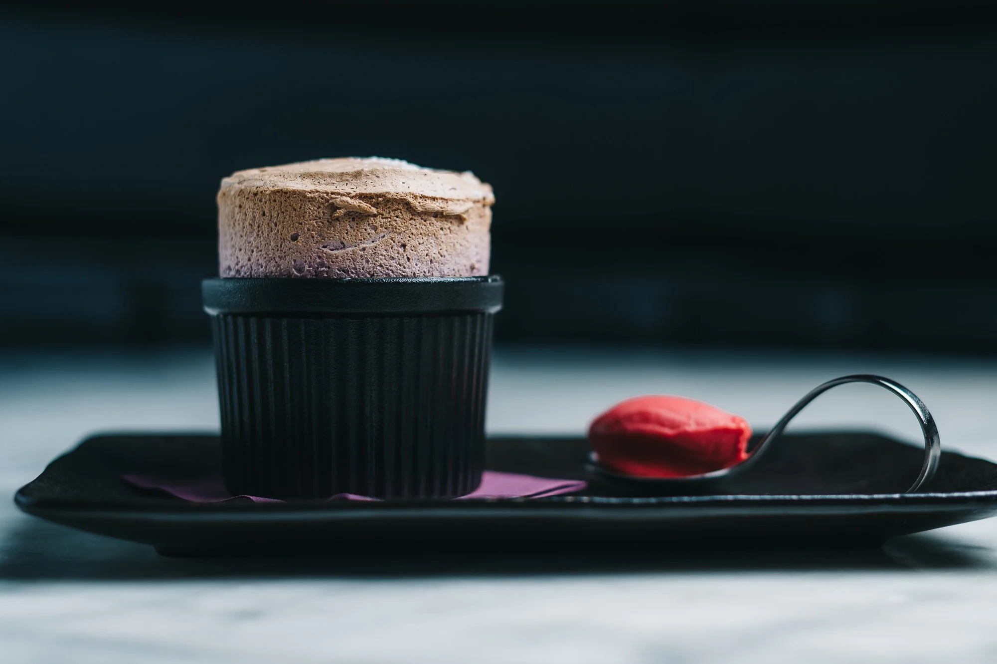 Chocolate souffle in a black cup on a black rectangular plate with a red spoon on a dark surface