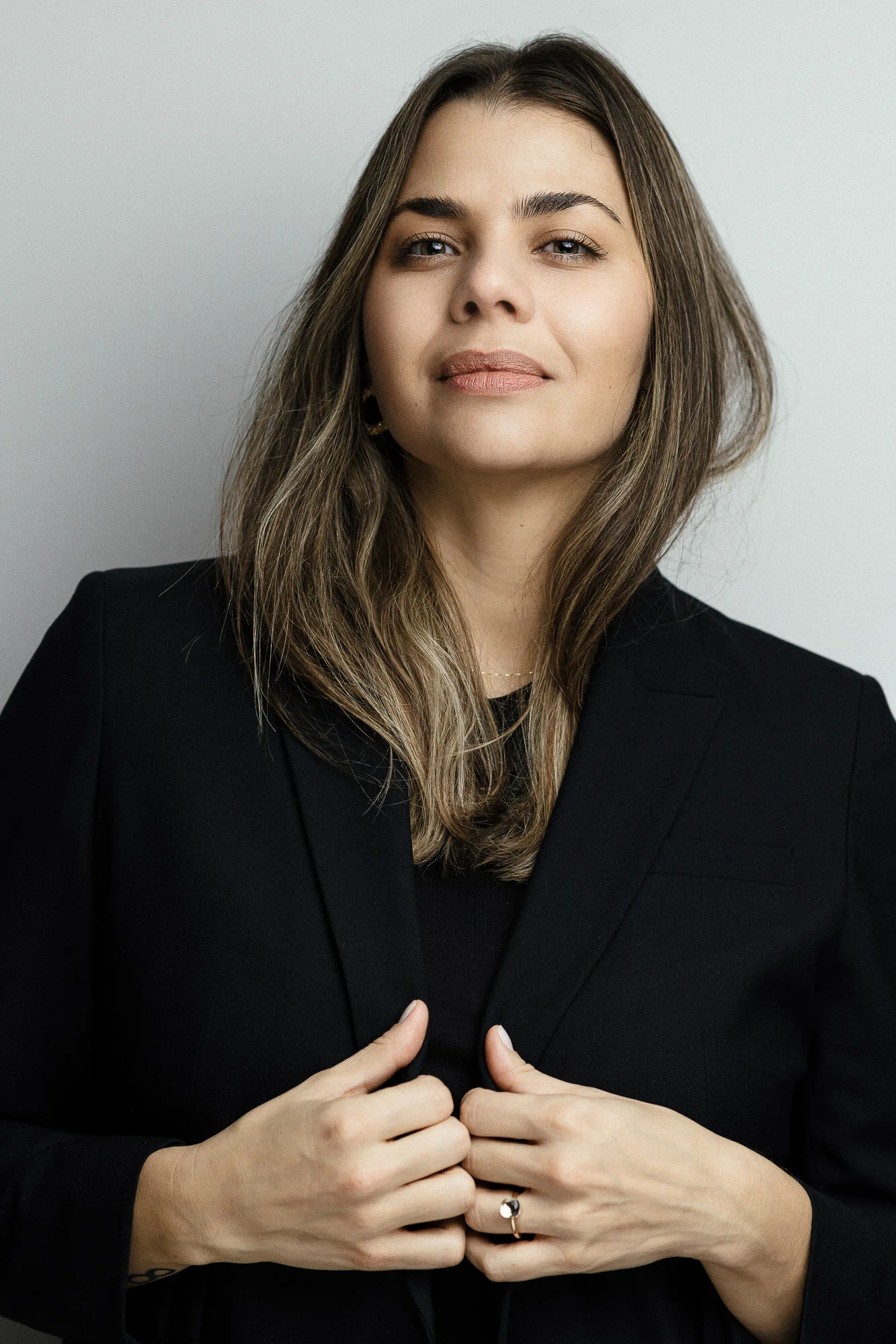 A woman with brown, wavy hair and light skin, wearing a black blazer and holding it with both hands, staring confidently at the camera against a plain light gray background.