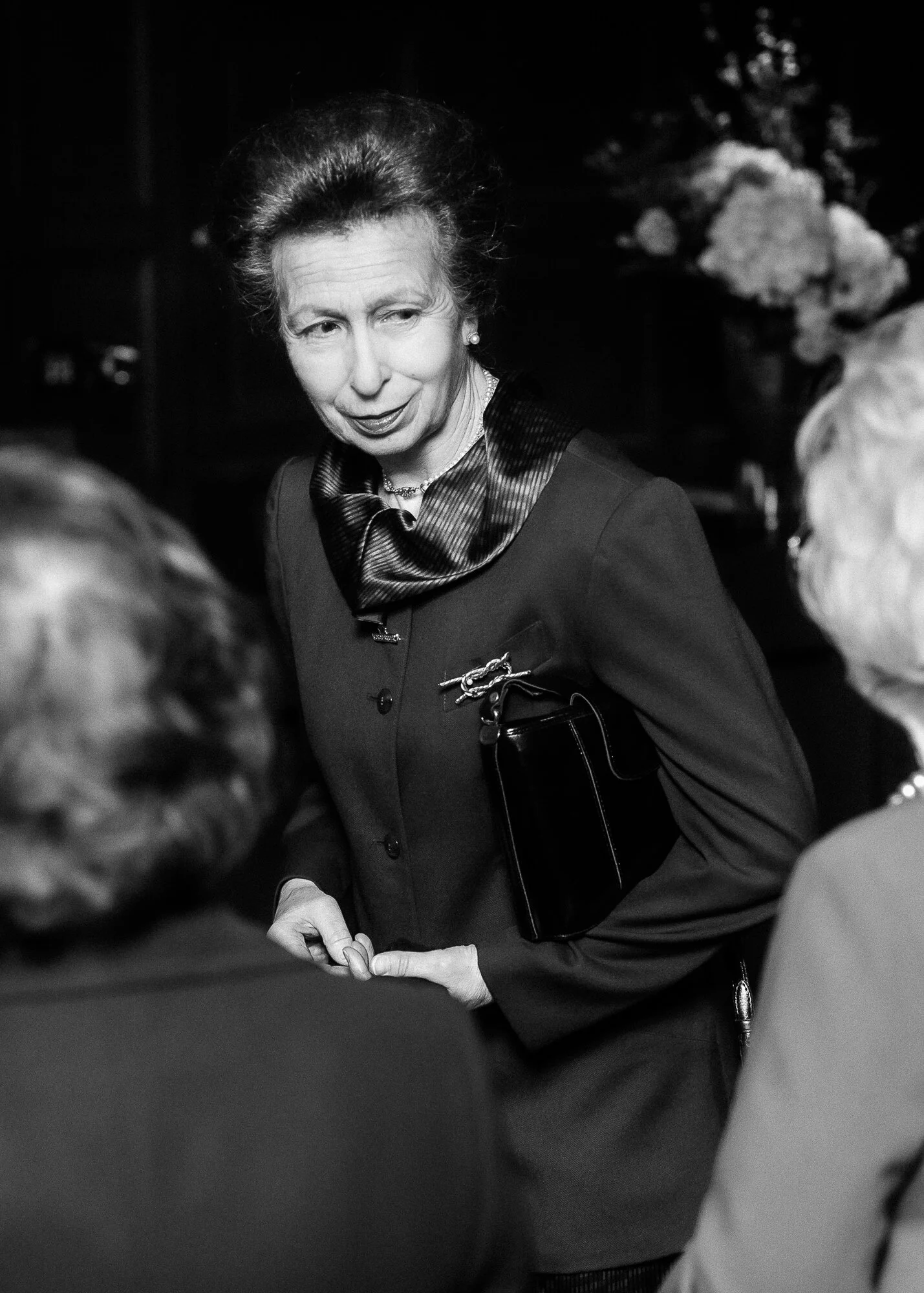 Black and white photo of a woman with styled hair, wearing a dark blazer and scarf, engaged in conversation with others, holding a purse.