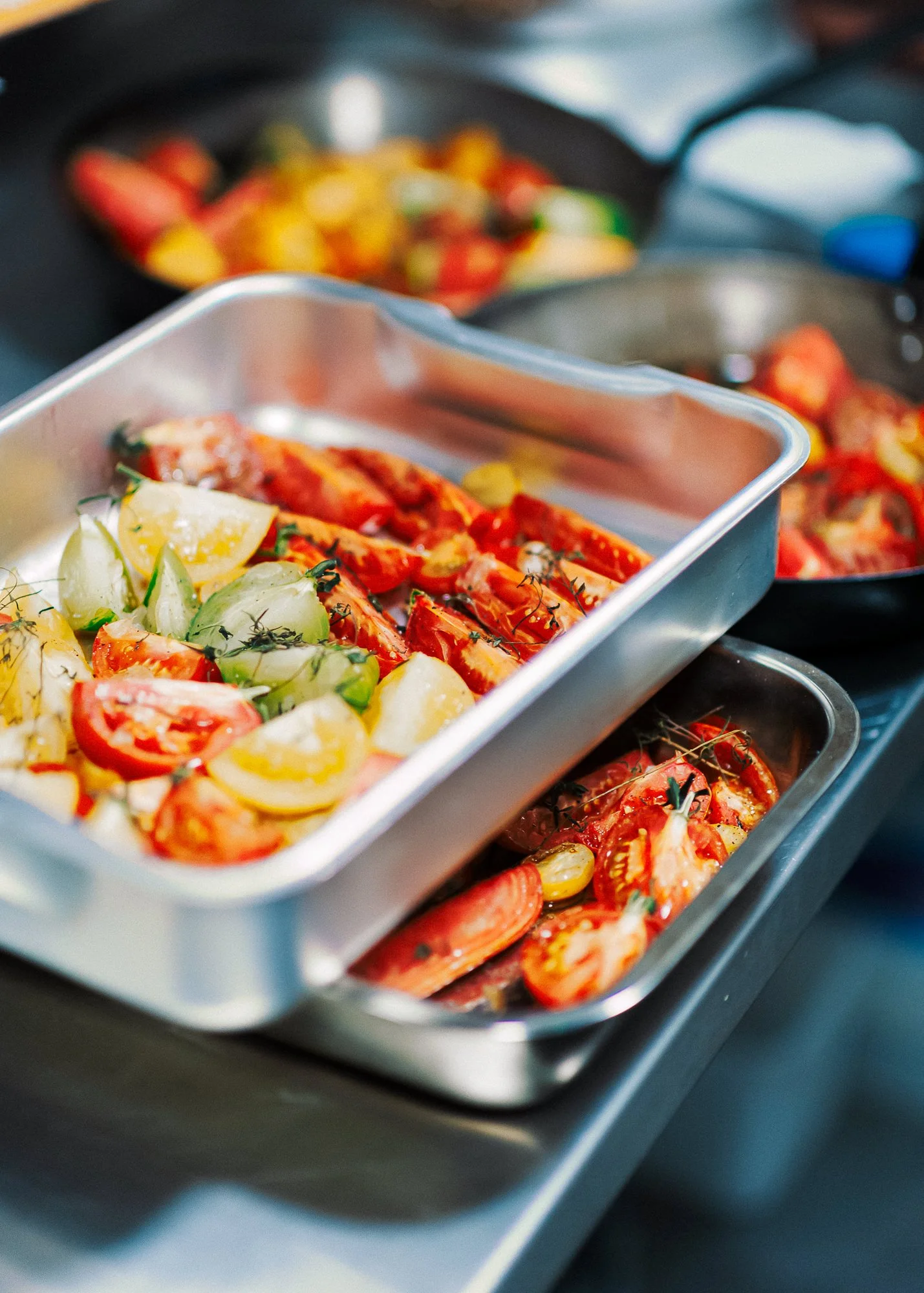 Three stainless steel containers filled with cooked tomatoes and vegetables, with other pans and kitchen tools in the background.